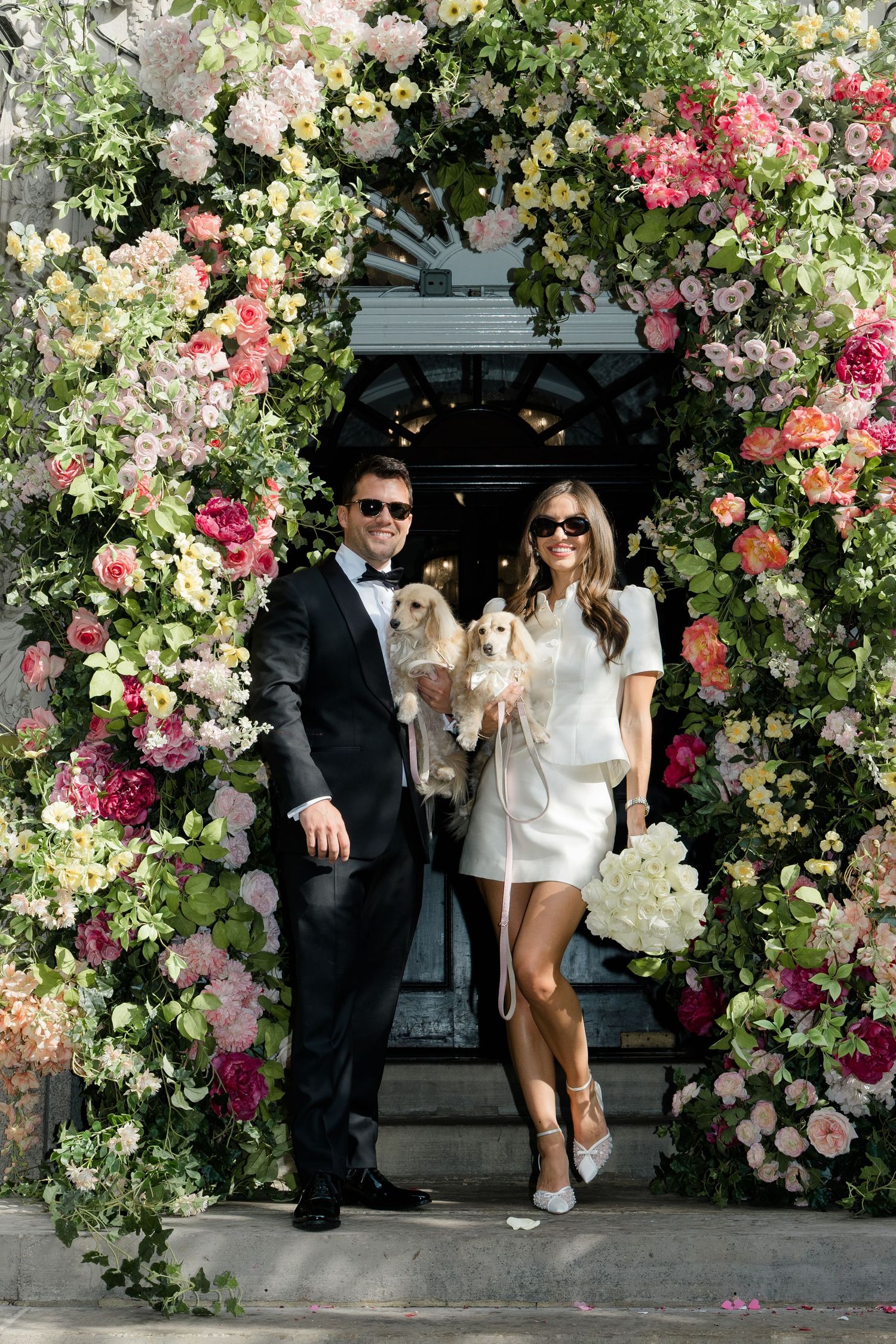 Couple in wedding attire posing with two dogs under a stunning floral arch adorned with pink roses and greenery.
