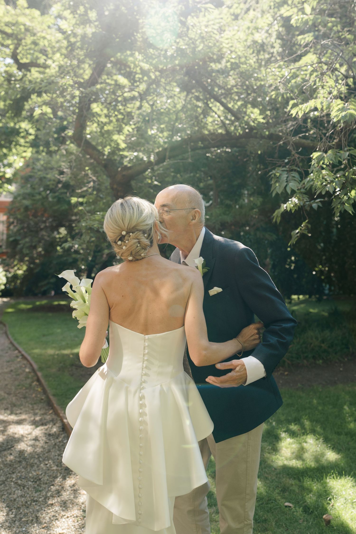 Bride in strapless white gown receives kiss from older man in navy suit in sunlit garden setting.