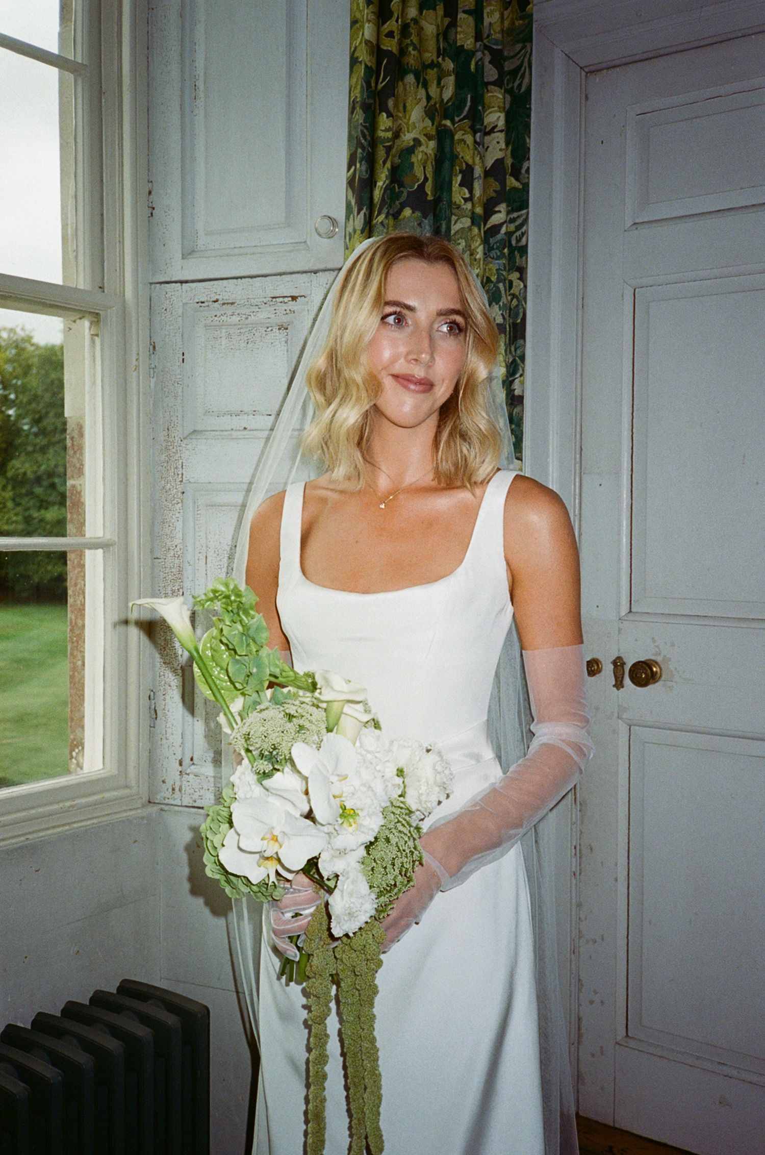 Bride in white gown holding green and white bouquet stands by vintage window in elegant venue.