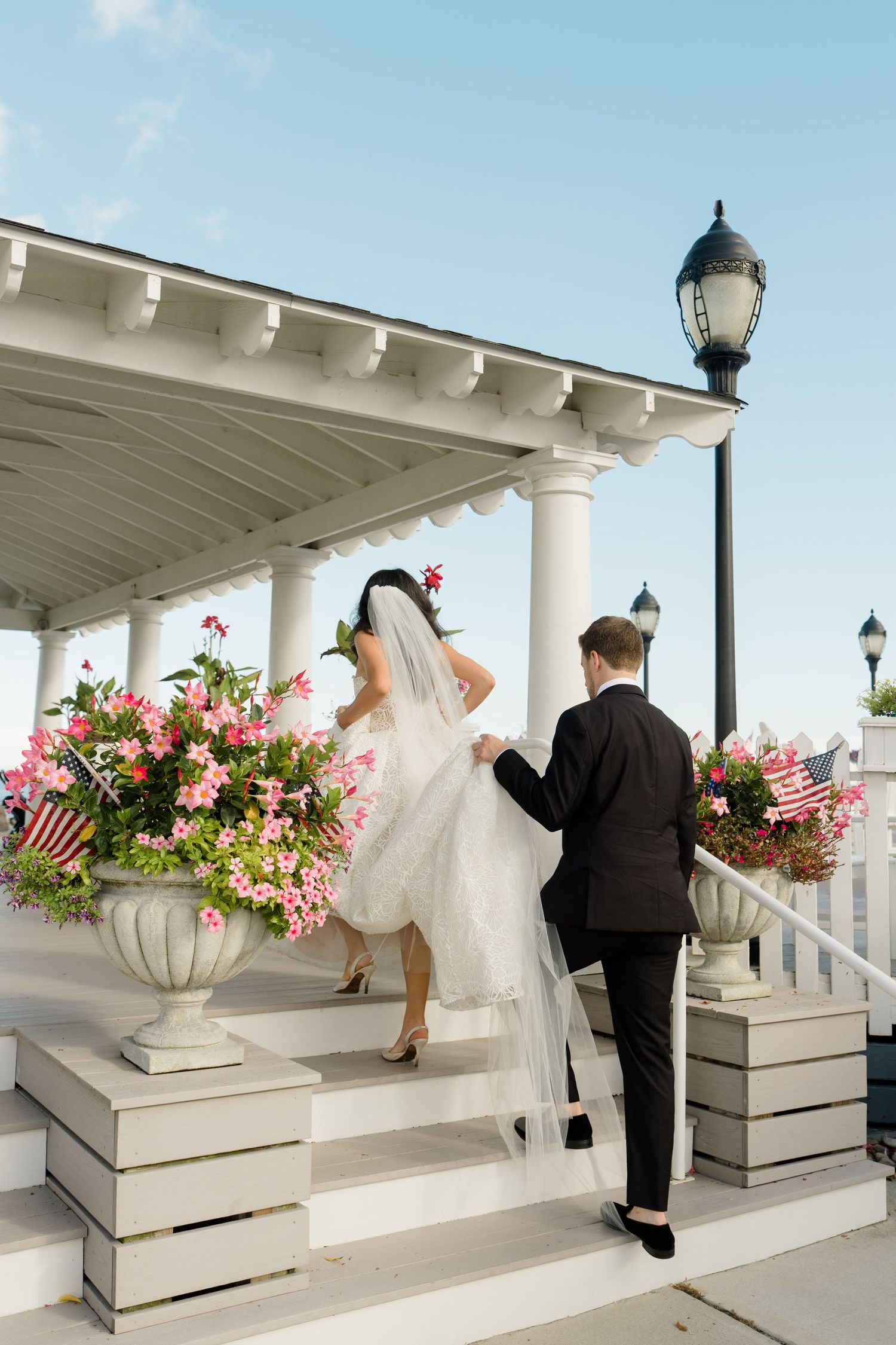 Bride and groom ascending steps of elegant white gazebo adorned with pink flowers and American flags on their wedding day.