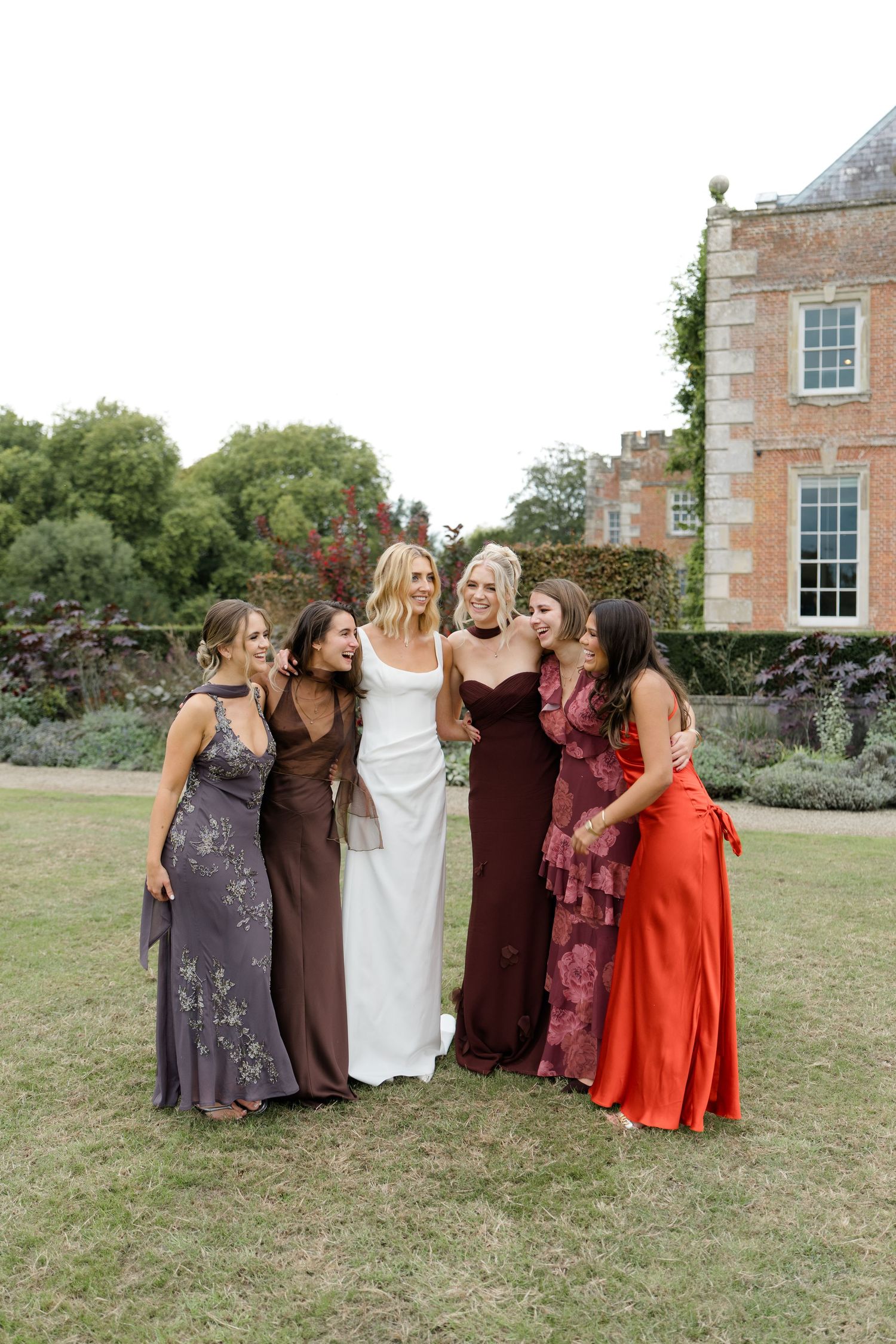 Bride in white gown surrounded by five bridesmaids in colorful autumn-toned dresses outside a historic manor.