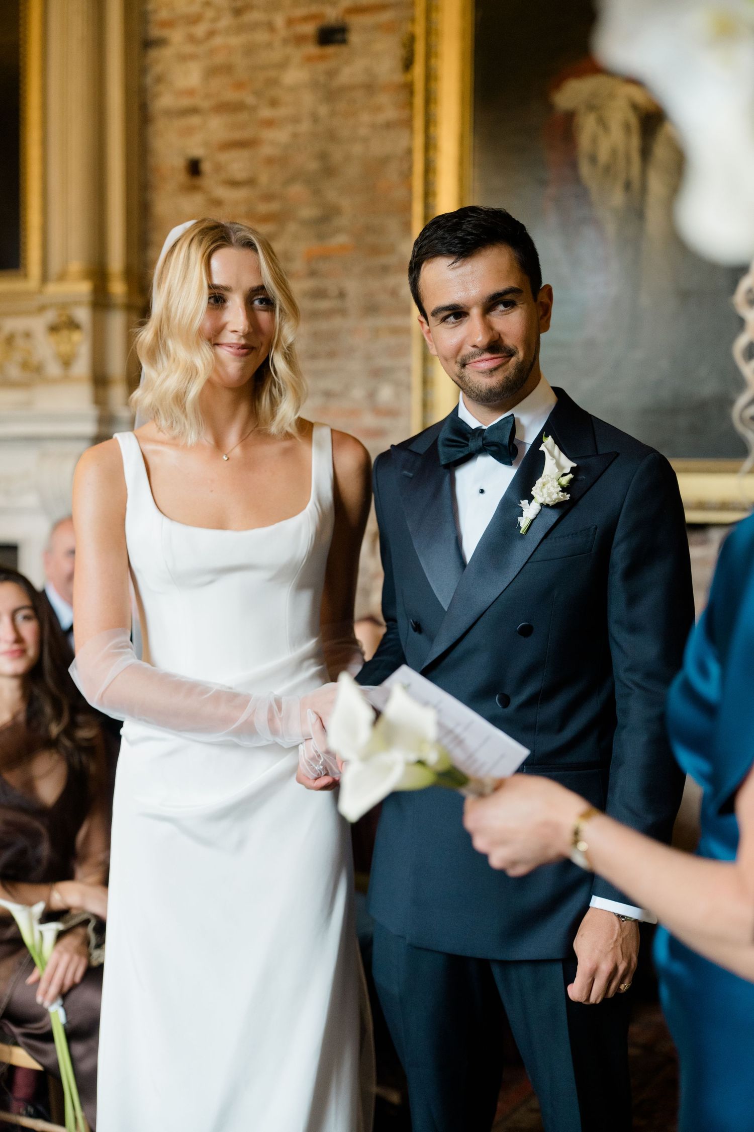 Bride and groom stand together during wedding ceremony, bride in sleek white dress, groom in navy tuxedo.