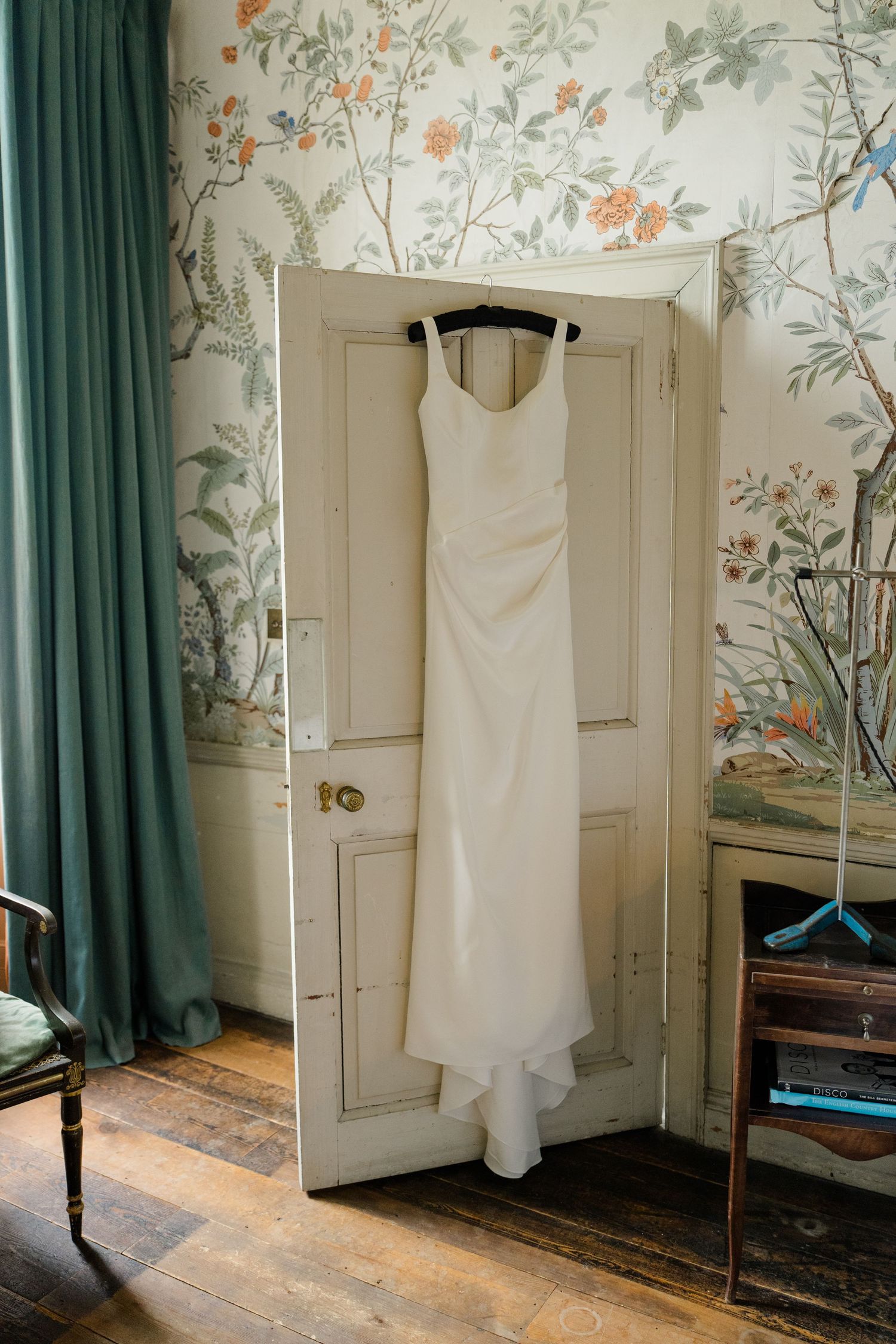 Simple white wedding dress hanging on a door against floral wallpaper in a vintage room.