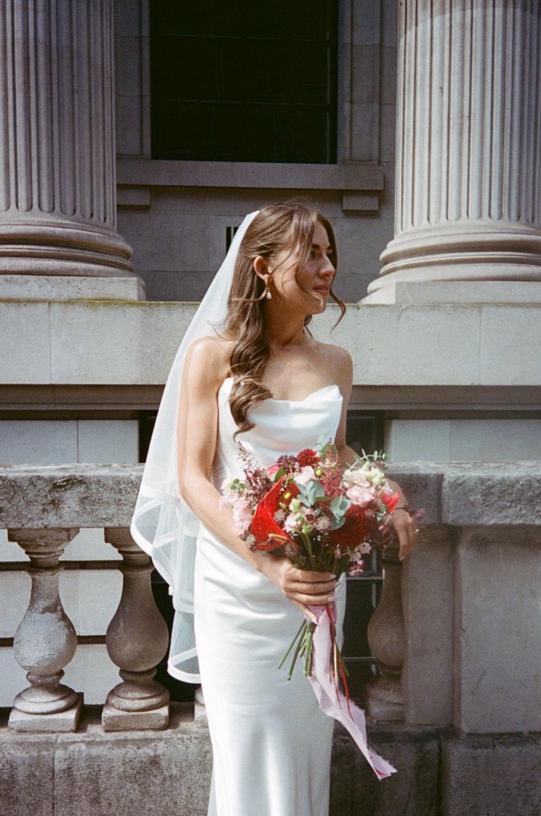 Bride in white strapless gown and veil holds red floral bouquet beside stone columns.