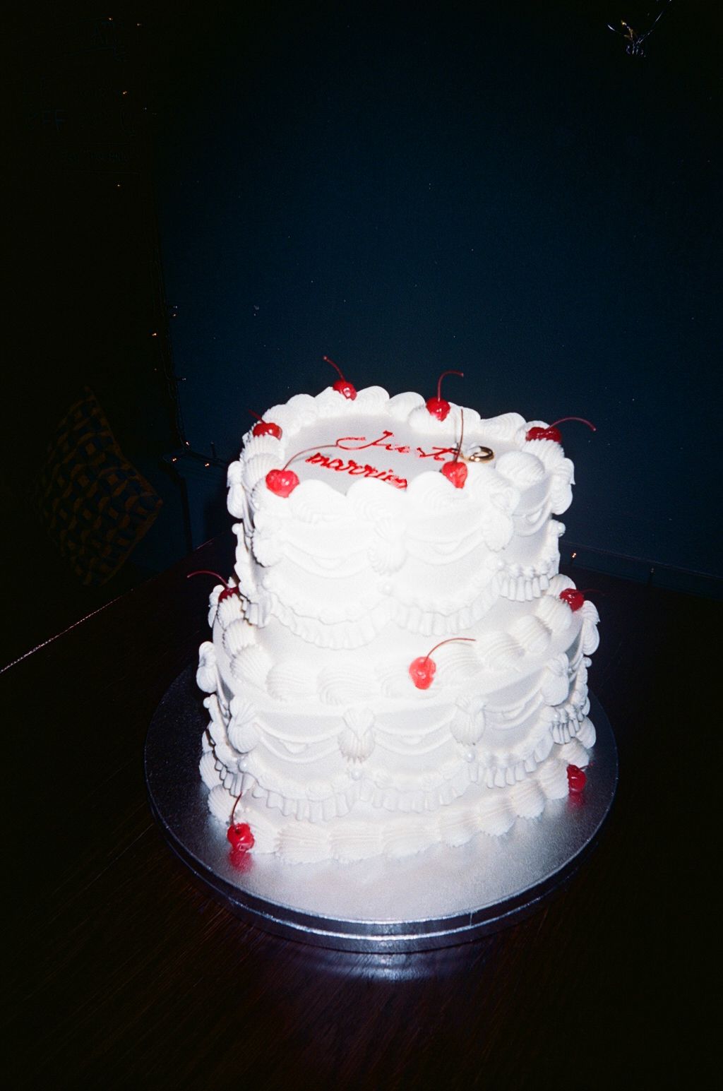 Two-tier white ruffled wedding cake decorated with red cherries on dark background.