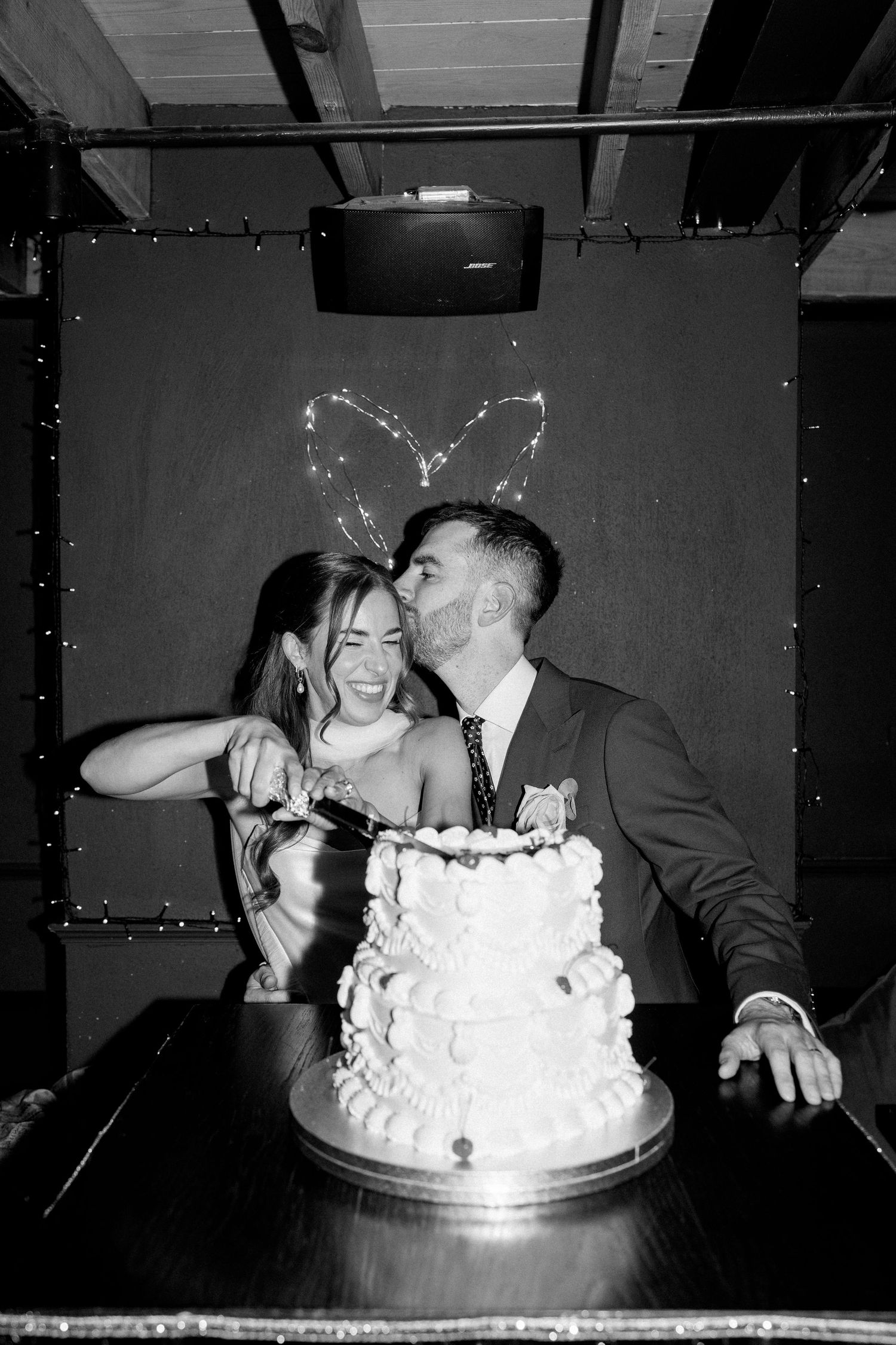 Black and white photo of couple cutting tiered wedding cake with heart light display.