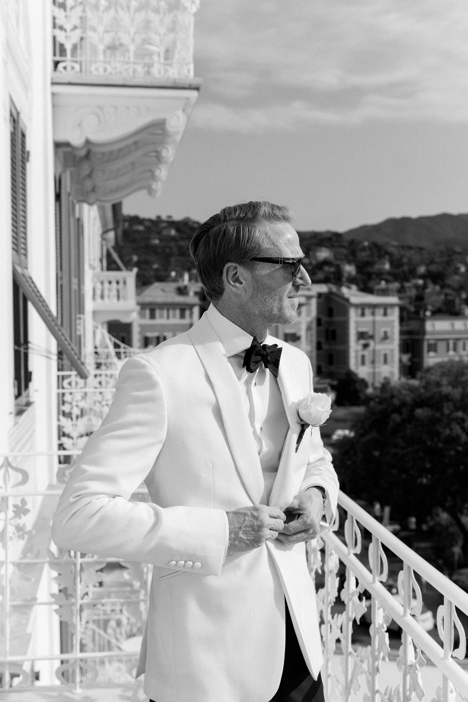 Groom in white tuxedo with boutonniere poses on ornate balcony overlooking Mediterranean town.
