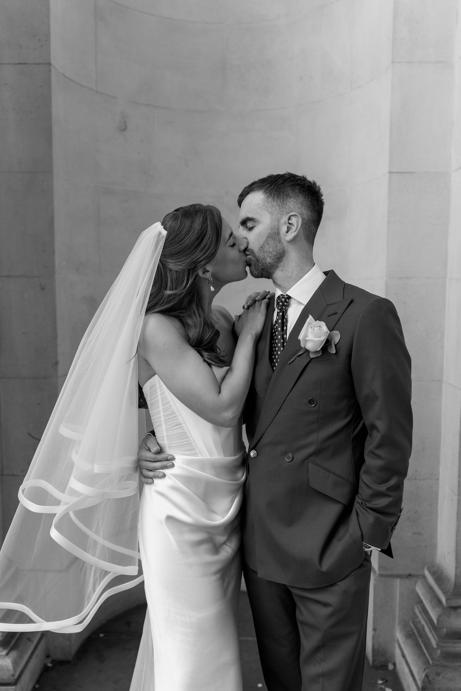 Black and white portrait of bride and groom sharing romantic kiss against stone column backdrop.