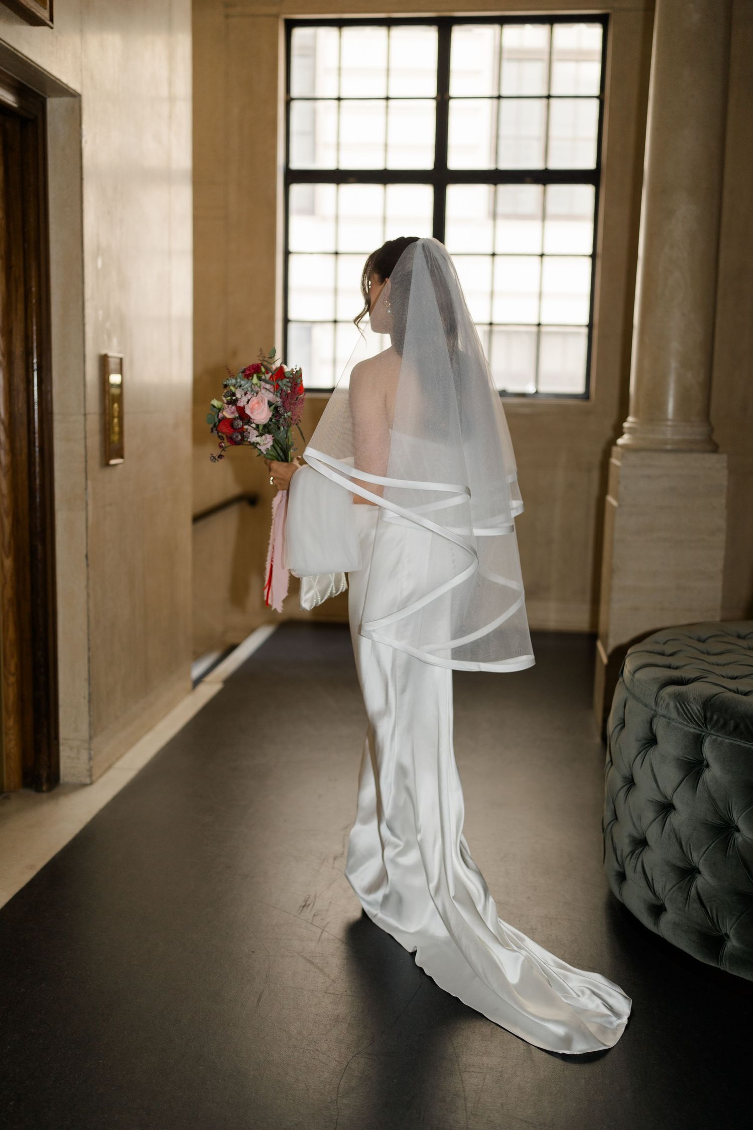 Bride in white satin gown and veil holds floral bouquet, standing by window in elegant indoor venue.