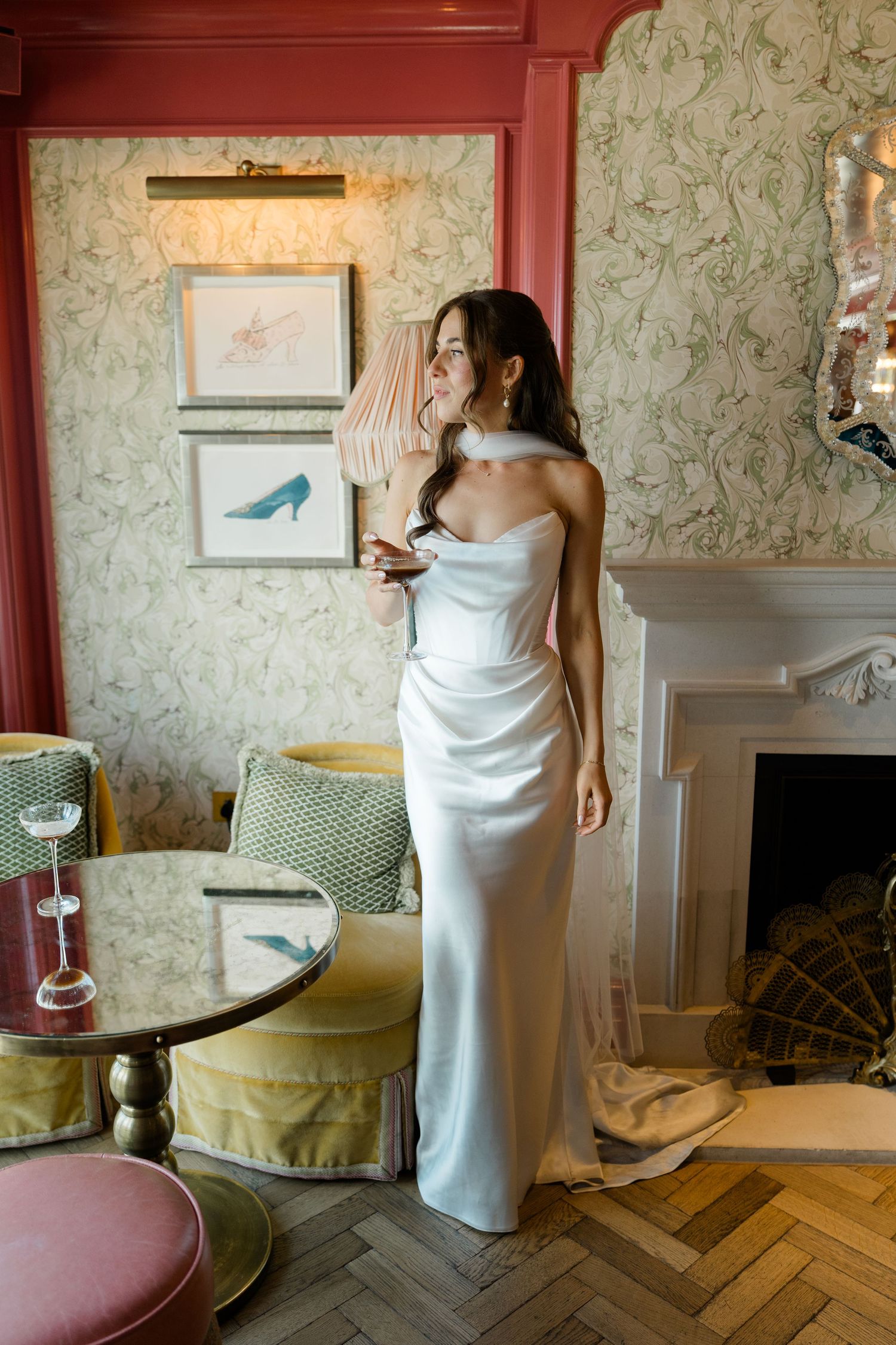 Elegant bride in strapless satin gown poses by fireplace in ornate floral wallpapered room.