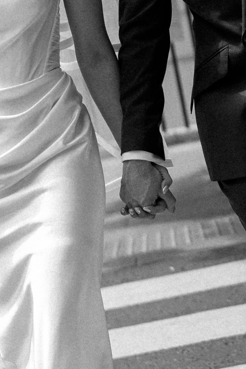 Black and white close-up of newlyweds holding hands while crossing street in wedding attire.
