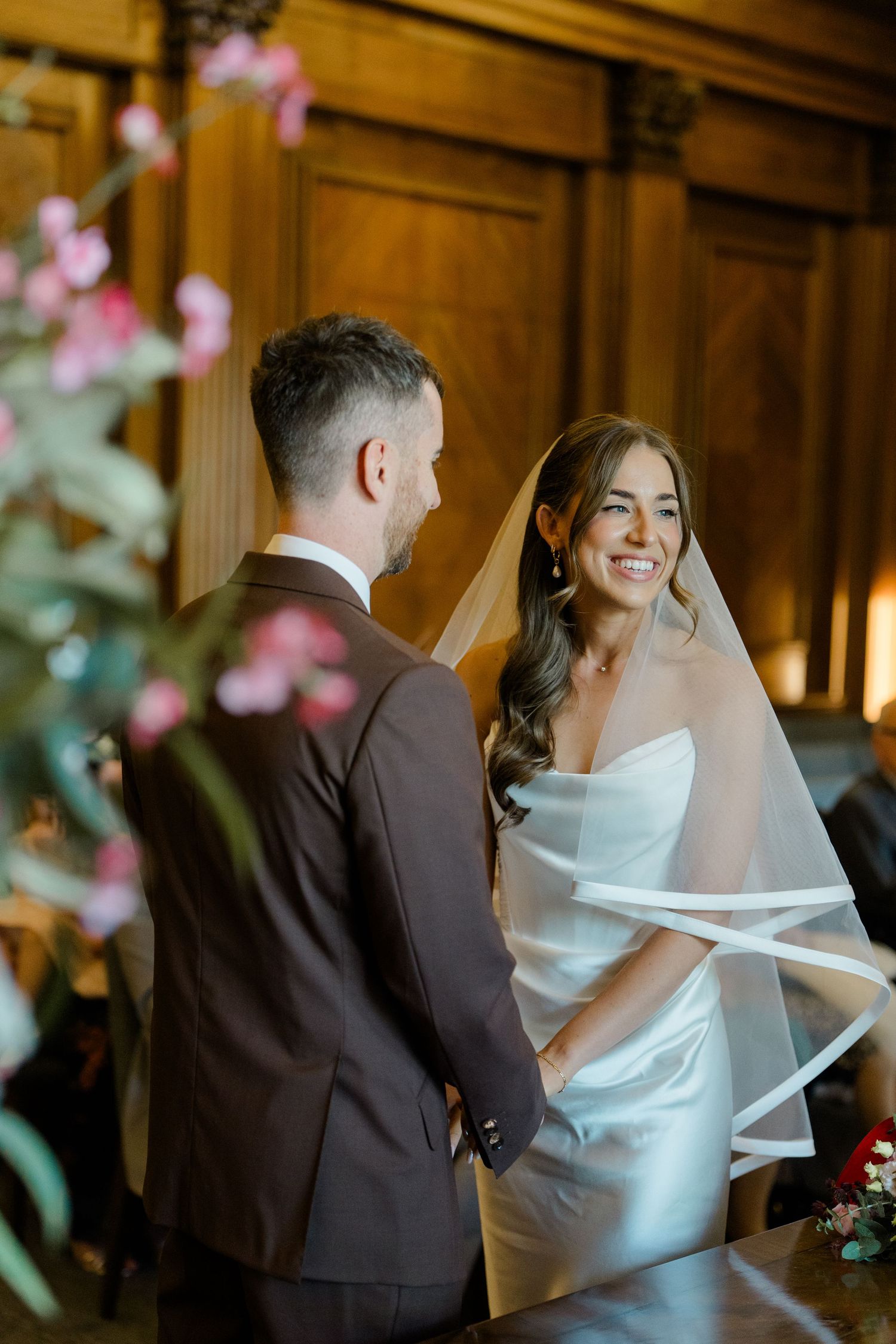 Bride smiling joyfully at groom during wedding ceremony in elegant wood-paneled room with floral decorations.