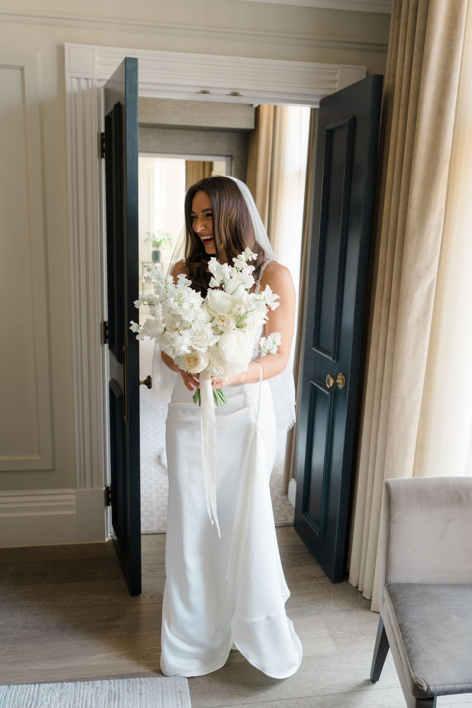Elegant bride in white gown and veil holds large white floral bouquet in doorway of hotel suite.