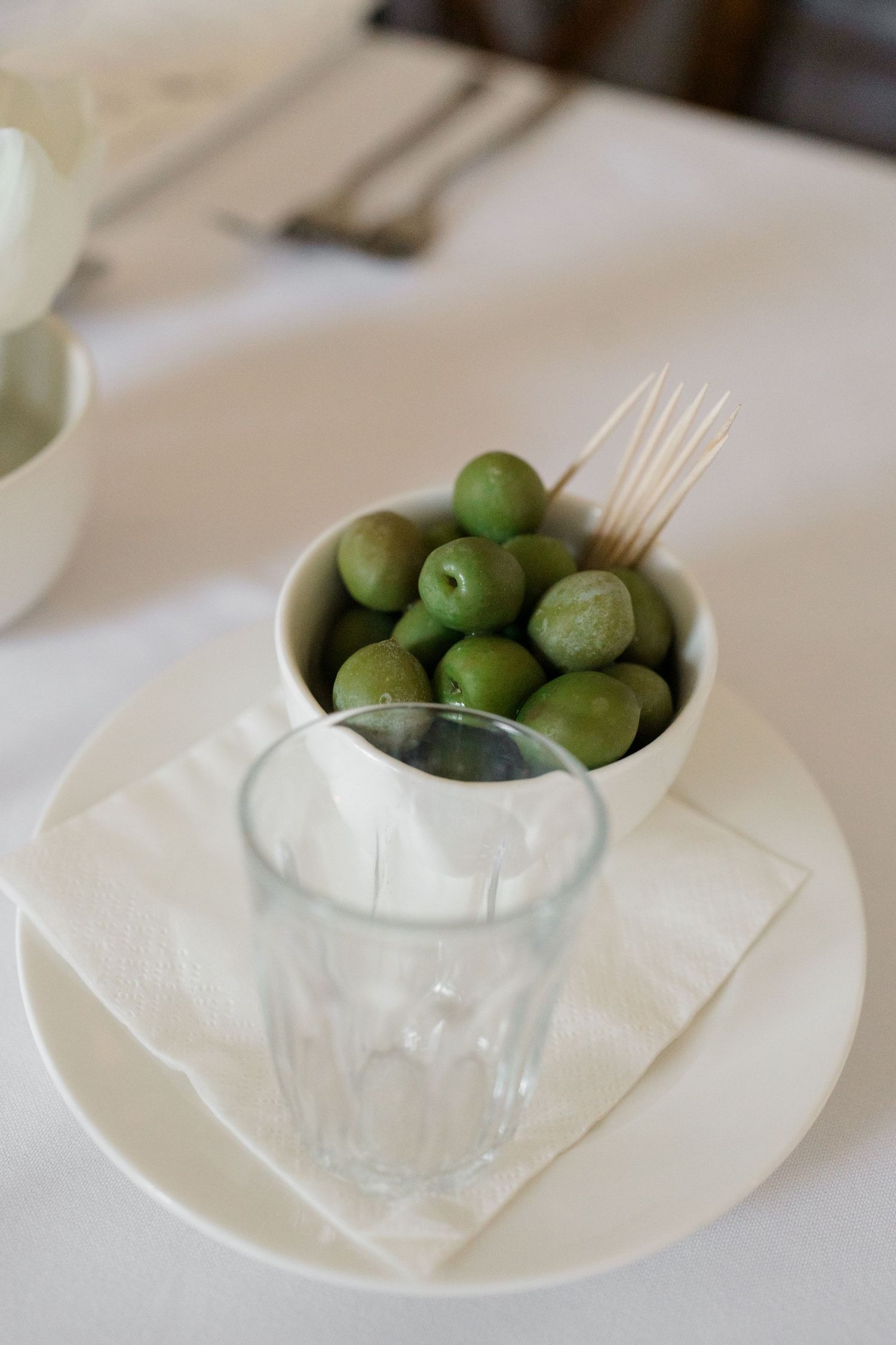 Bowl of green olives with toothpicks beside a glass on a white napkin at a wedding reception.