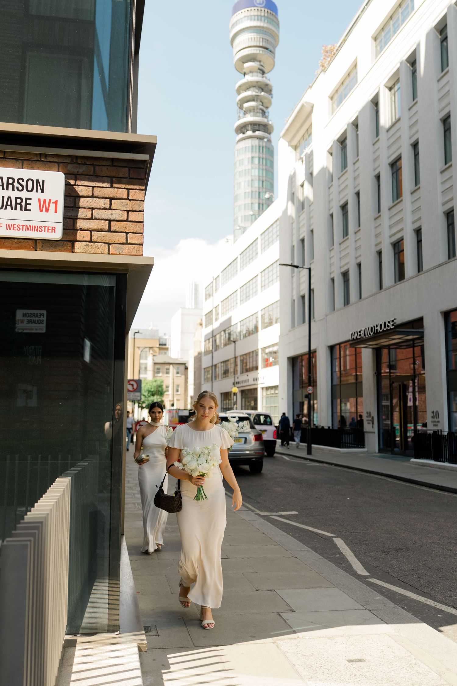 Bridesmaid walks along a London street near BT Tower holding white flowers on a sunny wedding day.
