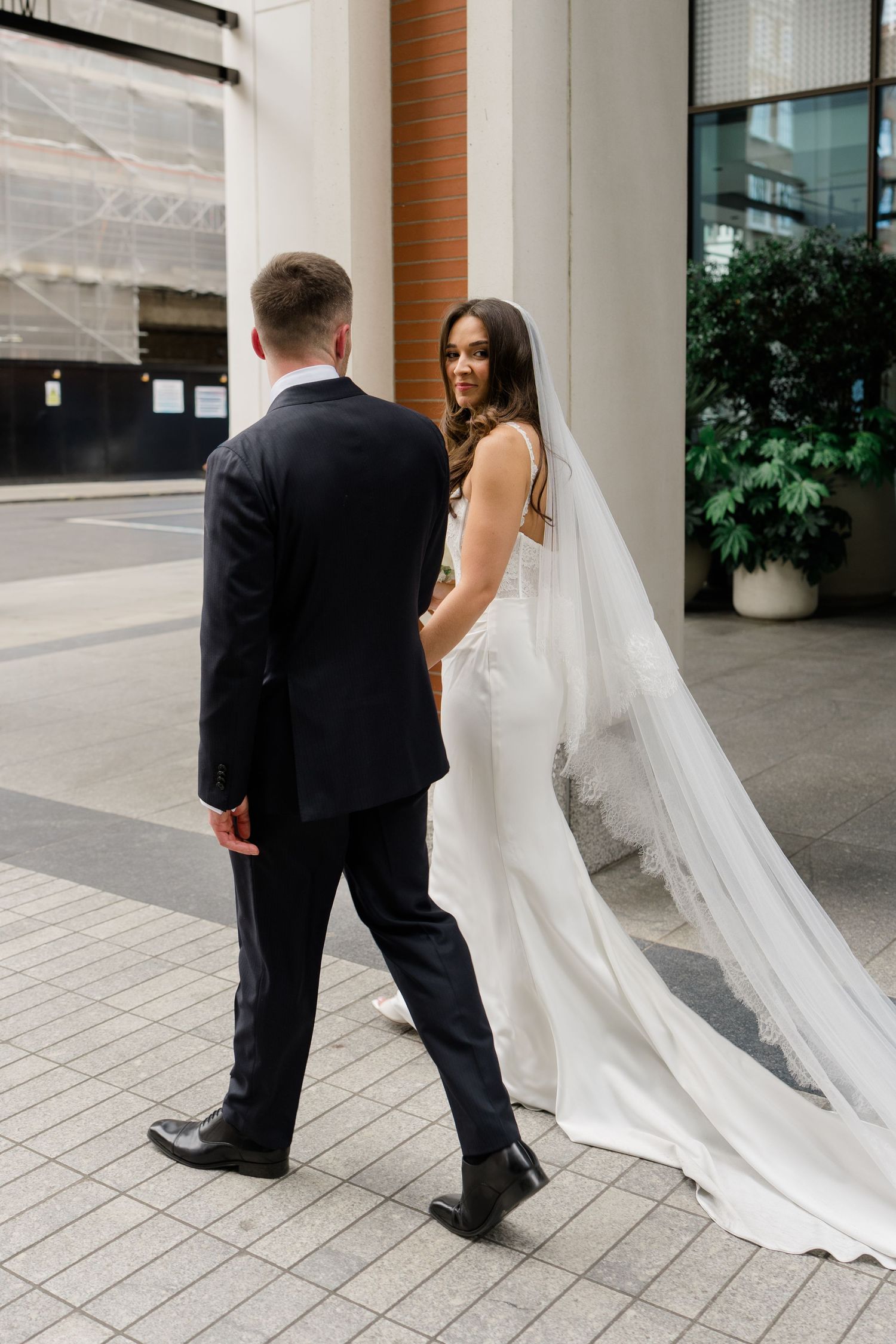 Bride in a stunning white gown with long veil walks hand in hand with groom on city street.