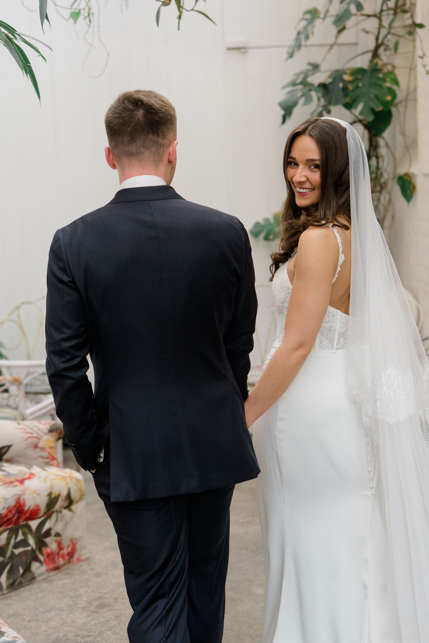 Bride in white gown and veil smiling over shoulder while holding grooms hand at wedding ceremony.