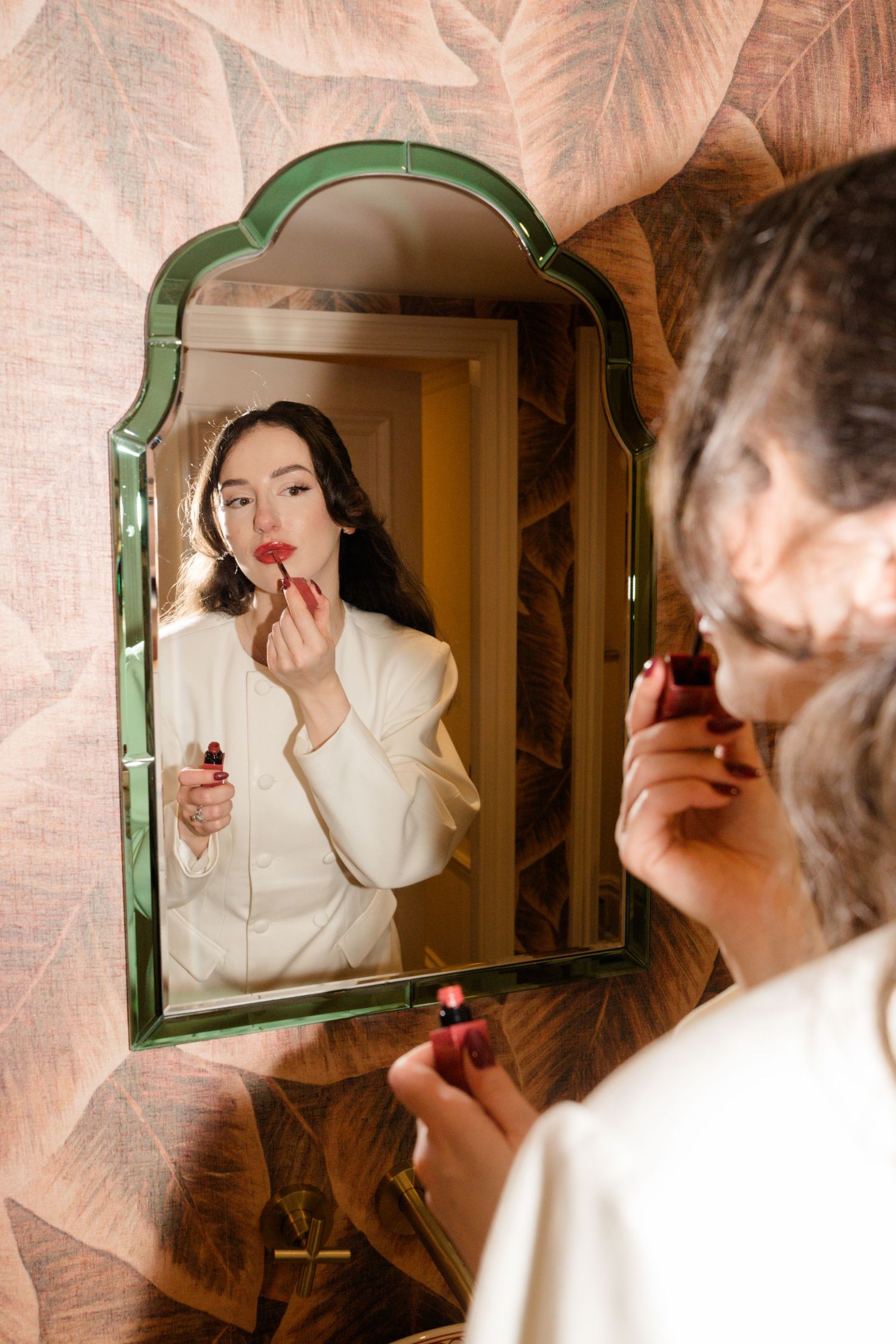Bride applies red lipstick while looking into ornate green-framed mirror with floral wallpaper backdrop.