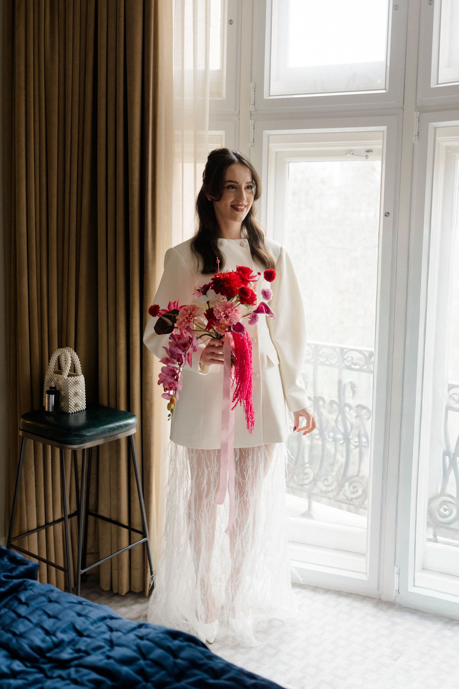 Bride in white mini dress with feather skirt holds vibrant red and pink bouquet by a bright window.