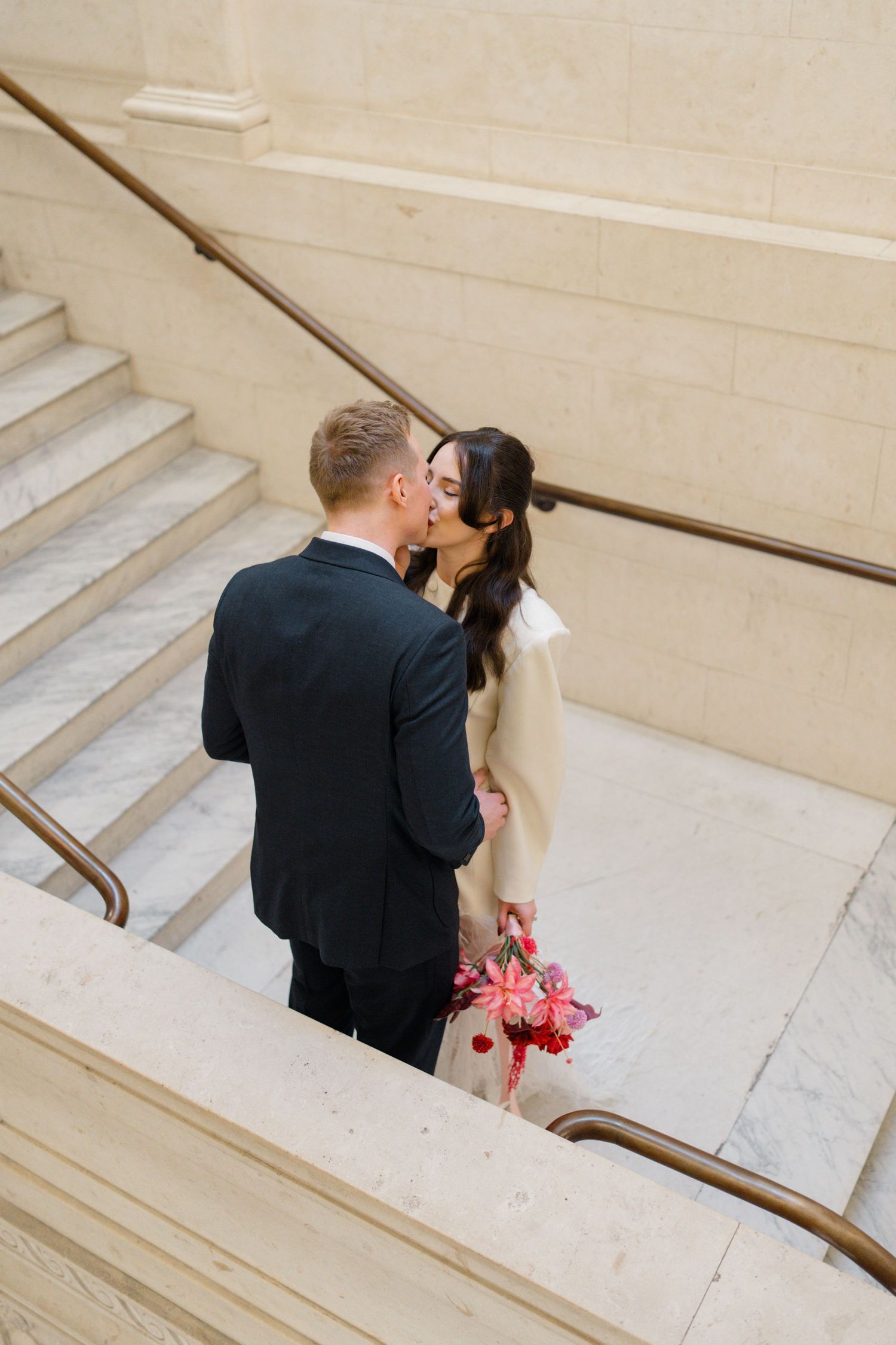 Newlyweds share a romantic kiss on marble staircase, bride holding red and pink bridal bouquet.