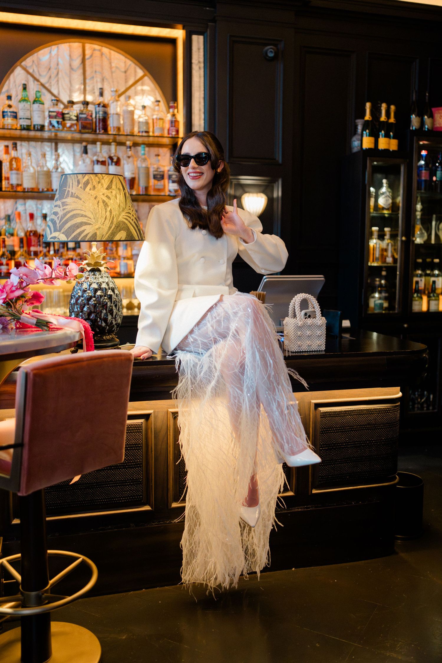 Stylish bride in white blazer and feather skirt waving at luxury hotel bar with pink bouquet.