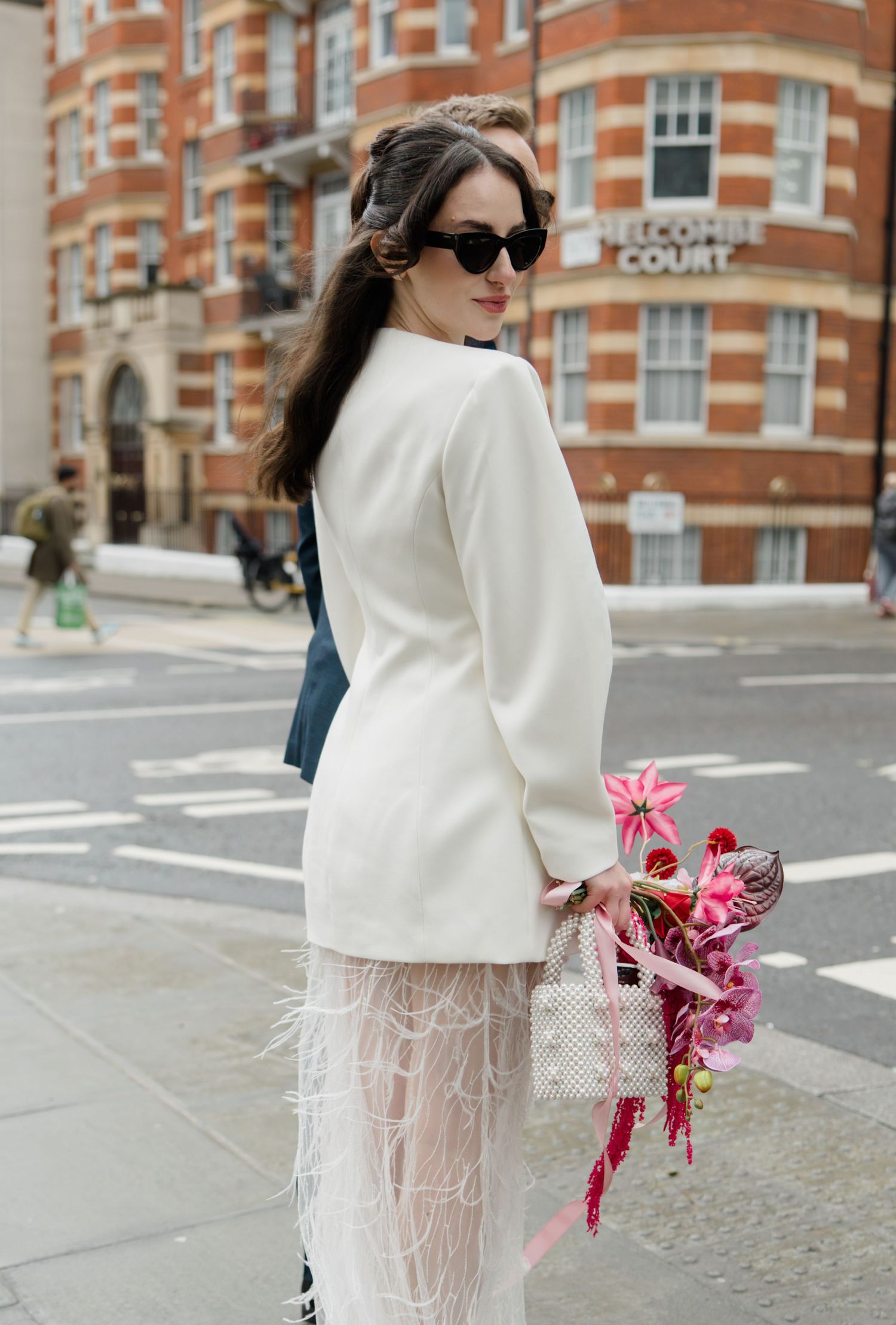 Stylish bride in white blazer and feather skirt wearing sunglasses holds colorful bouquet on London street.
