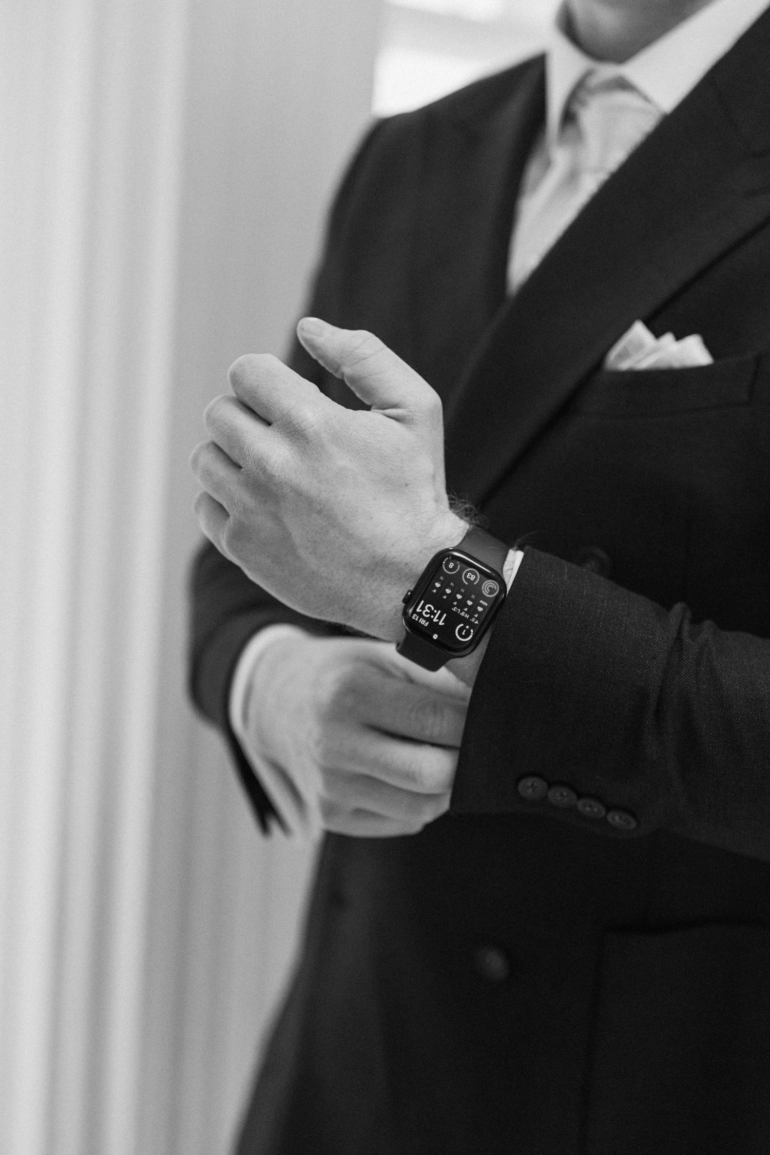 Groom in dark suit adjusting Apple Watch cufflinks, black and white wedding detail photo.