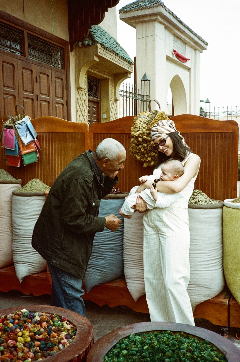 A woman in white holding a baby interacts with an elderly man near colorful spice sacks at a Moroccan market.