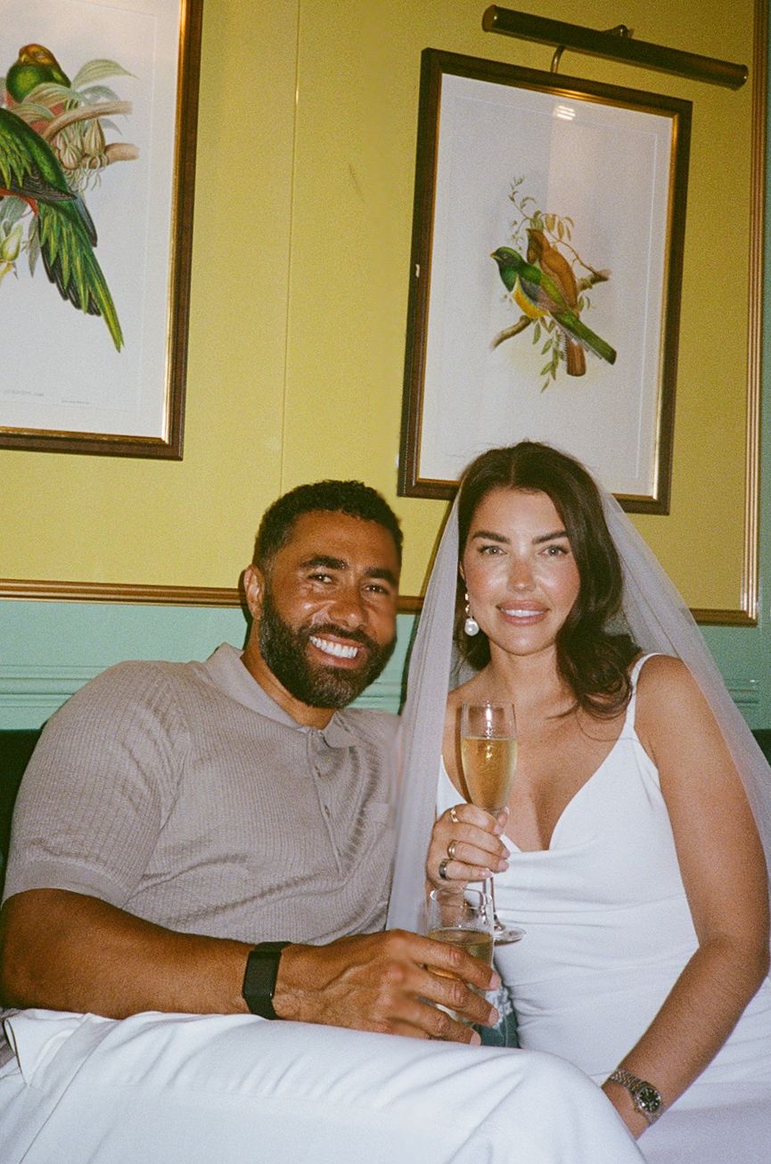 A smiling couple in wedding attire celebrates with champagne glasses in a green room with bird artwork.