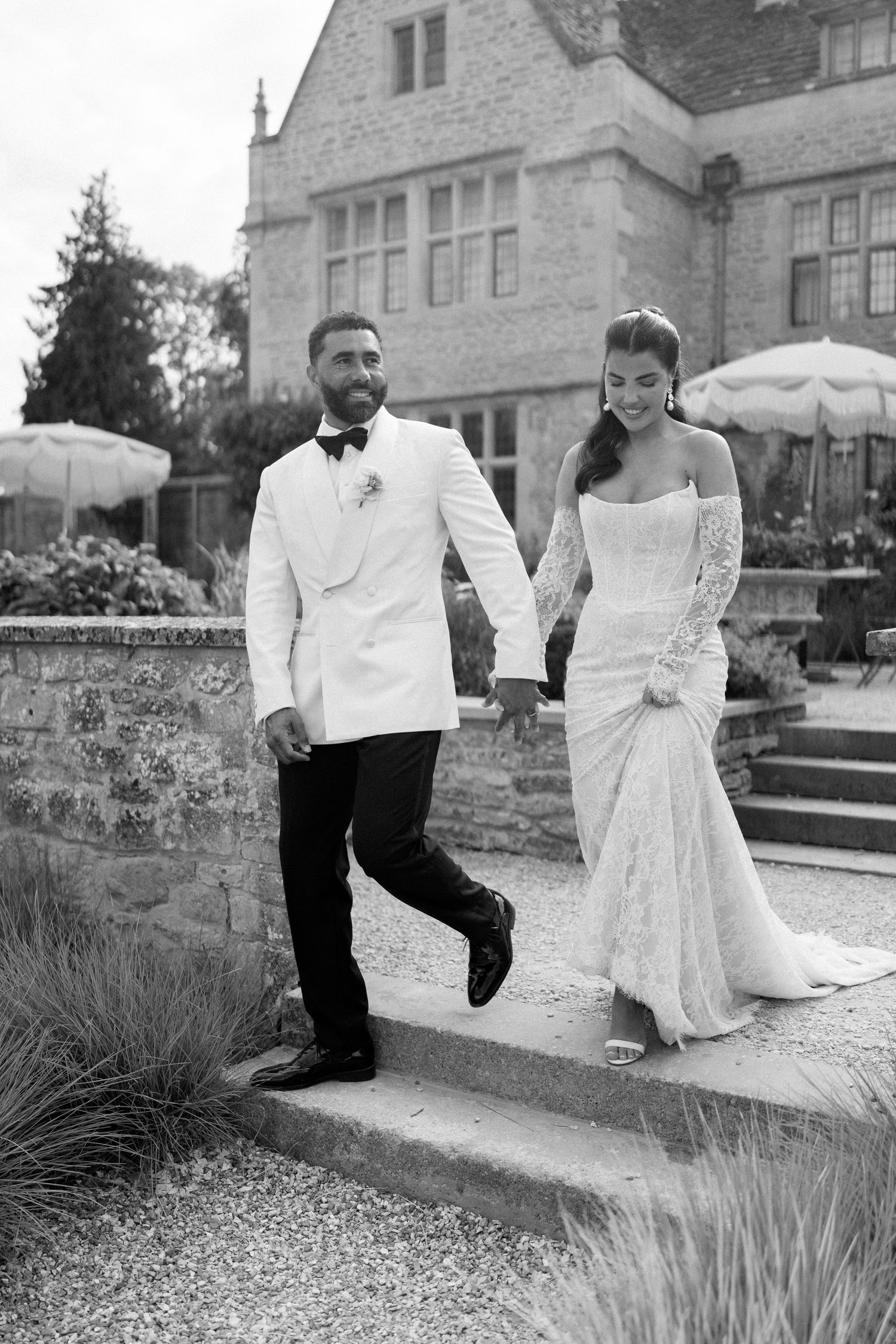 Black and white photo of bride and groom holding hands walking down steps outside historic manor house.