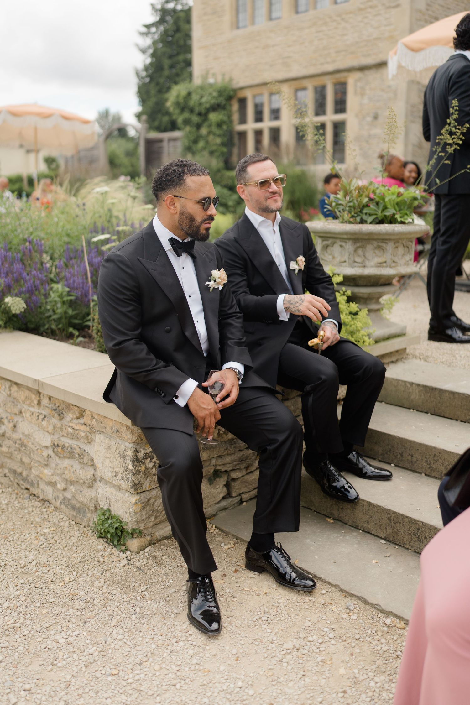 Two groomsmen in black tuxedos with boutonnieres sitting on stone steps at wedding.