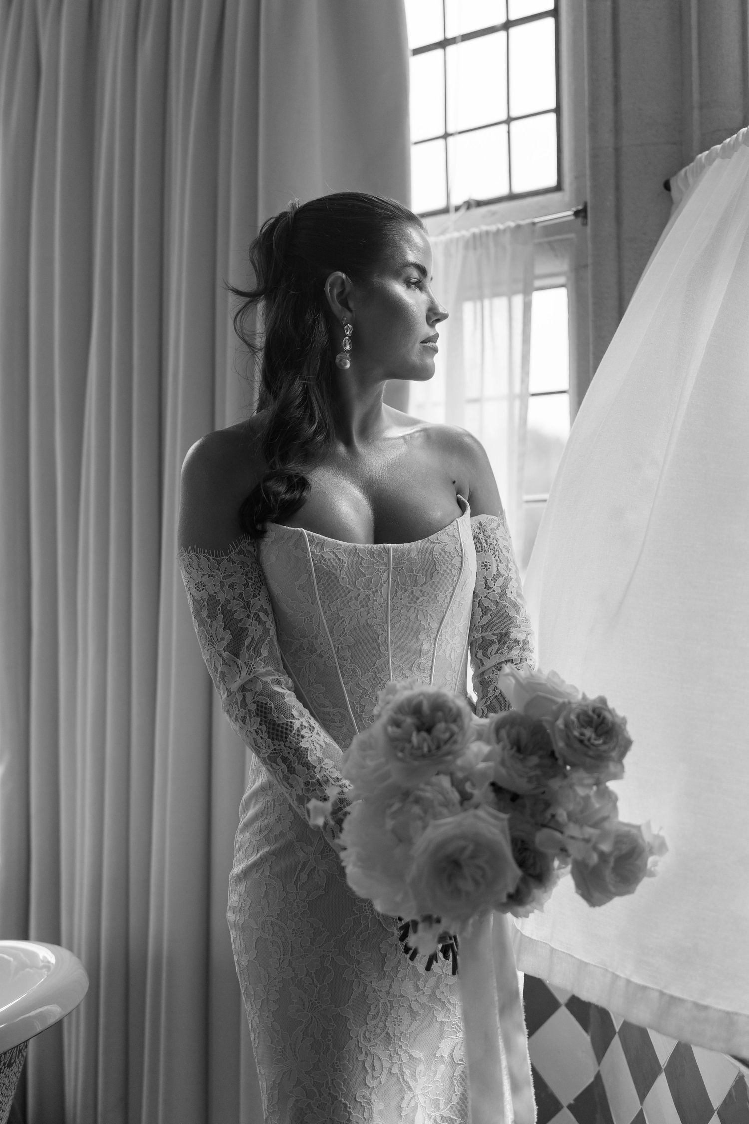Elegant bride in lace wedding gown holding peony bouquet by window in black and white portrait.