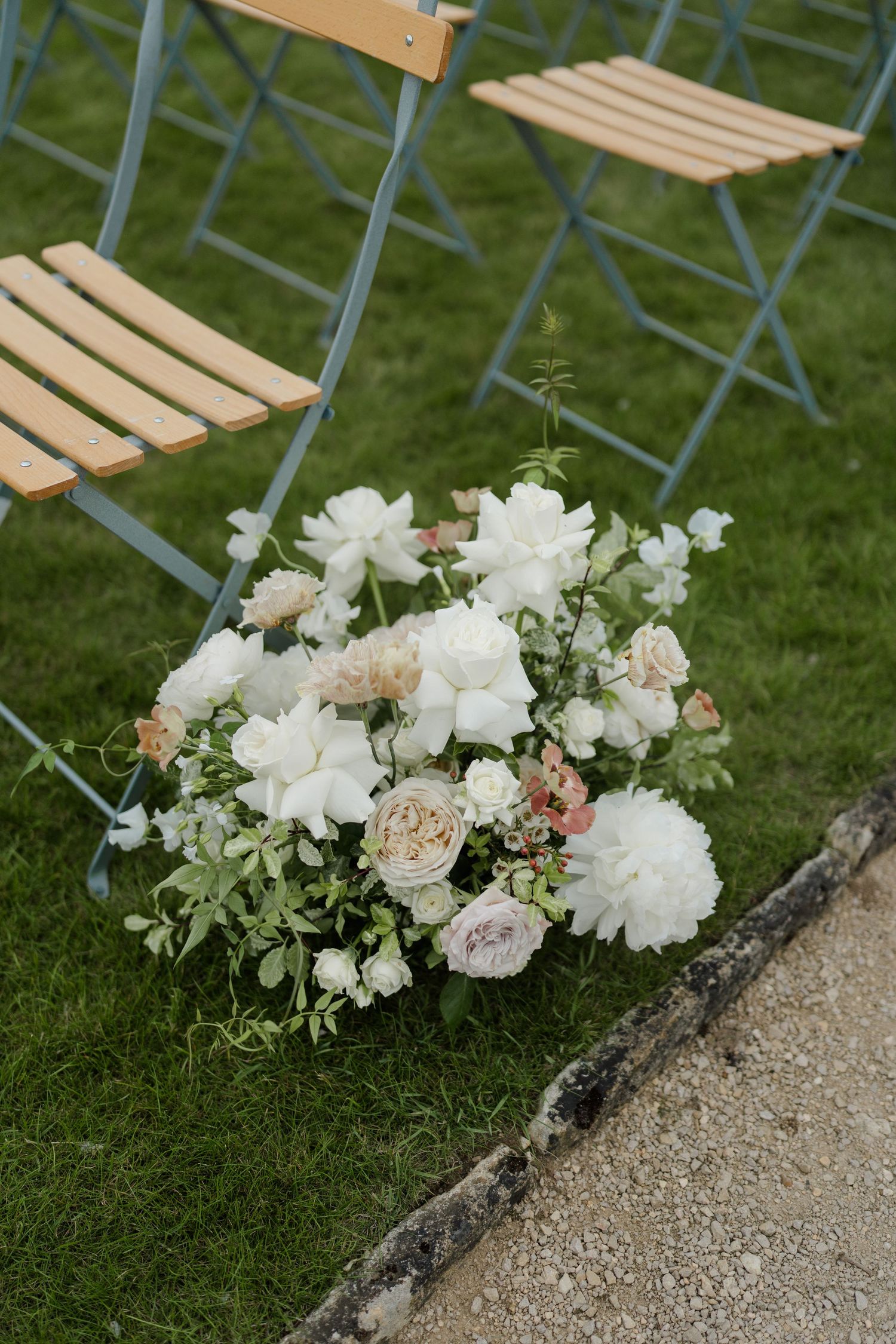 White and blush floral arrangement with peonies and roses beside wooden folding chairs at outdoor ceremony.