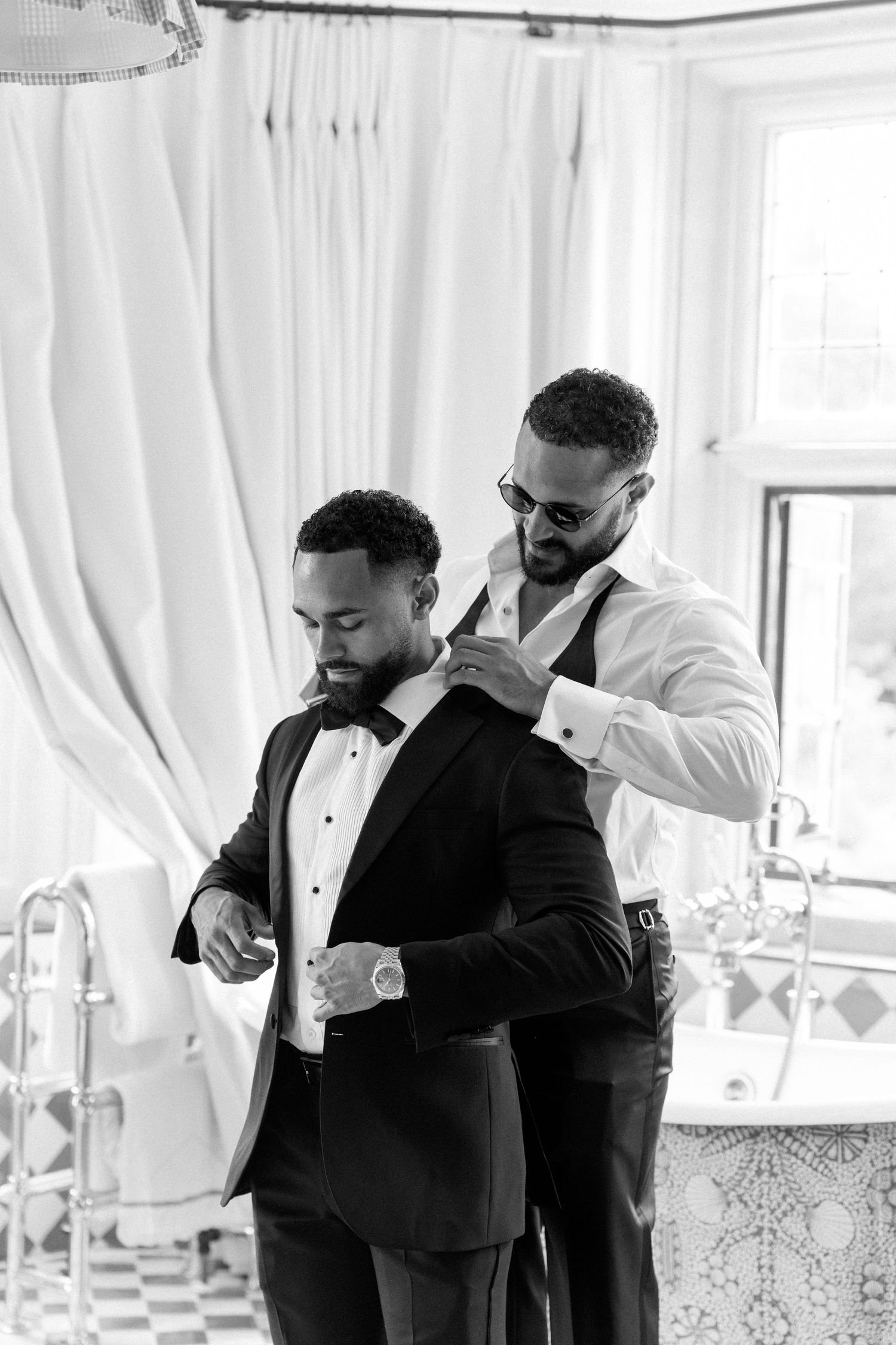 Black and white photo of a groomsman helping the groom adjust his tuxedo jacket.