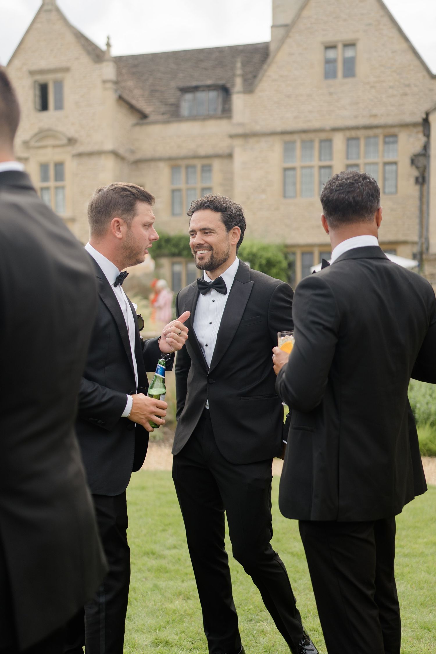 Three groomsmen in black tuxedos laugh and chat together on a lawn outside a stone manor wedding venue.