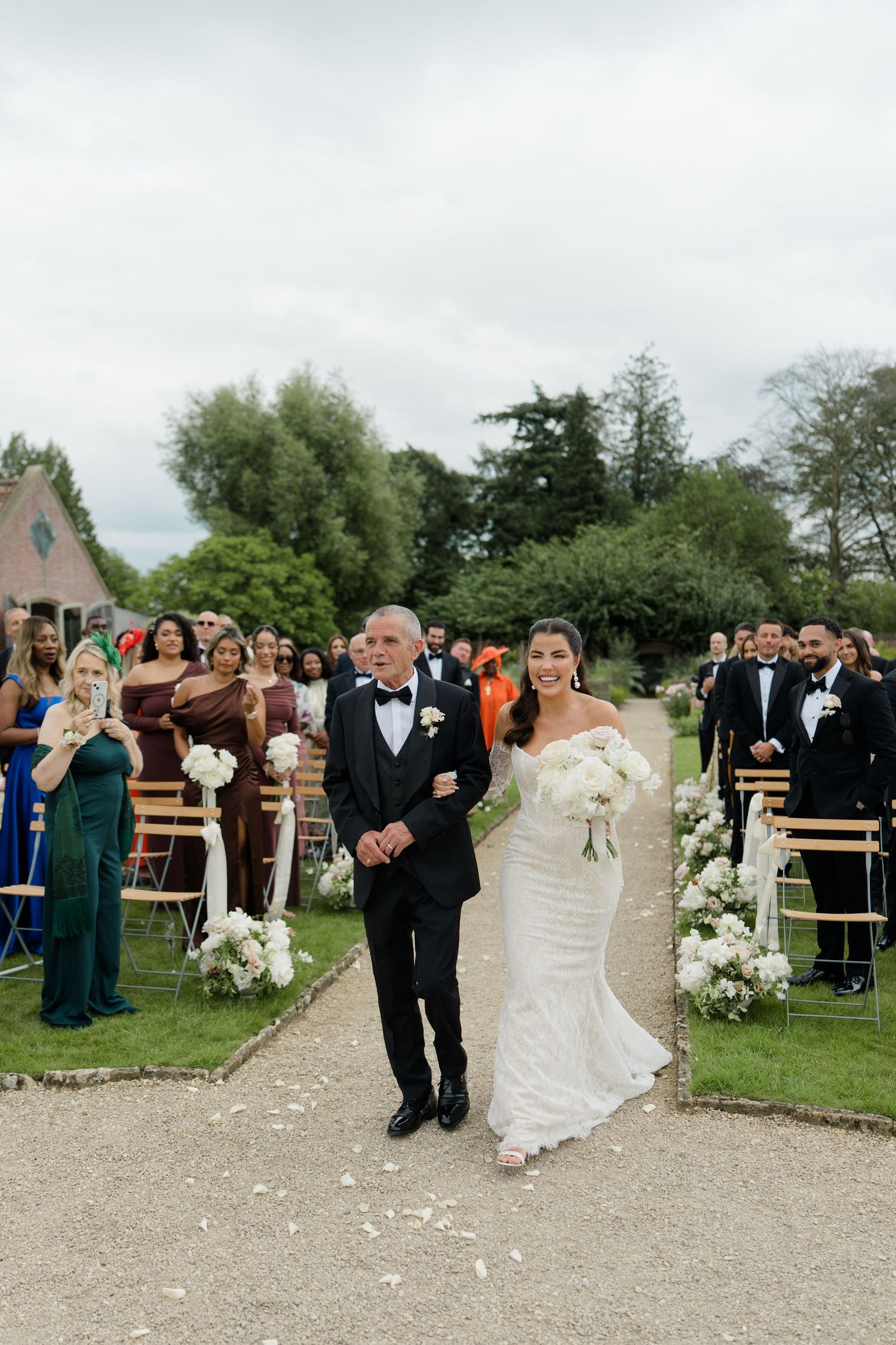Bride in fitted white gown walks down outdoor aisle with father during wedding ceremony.