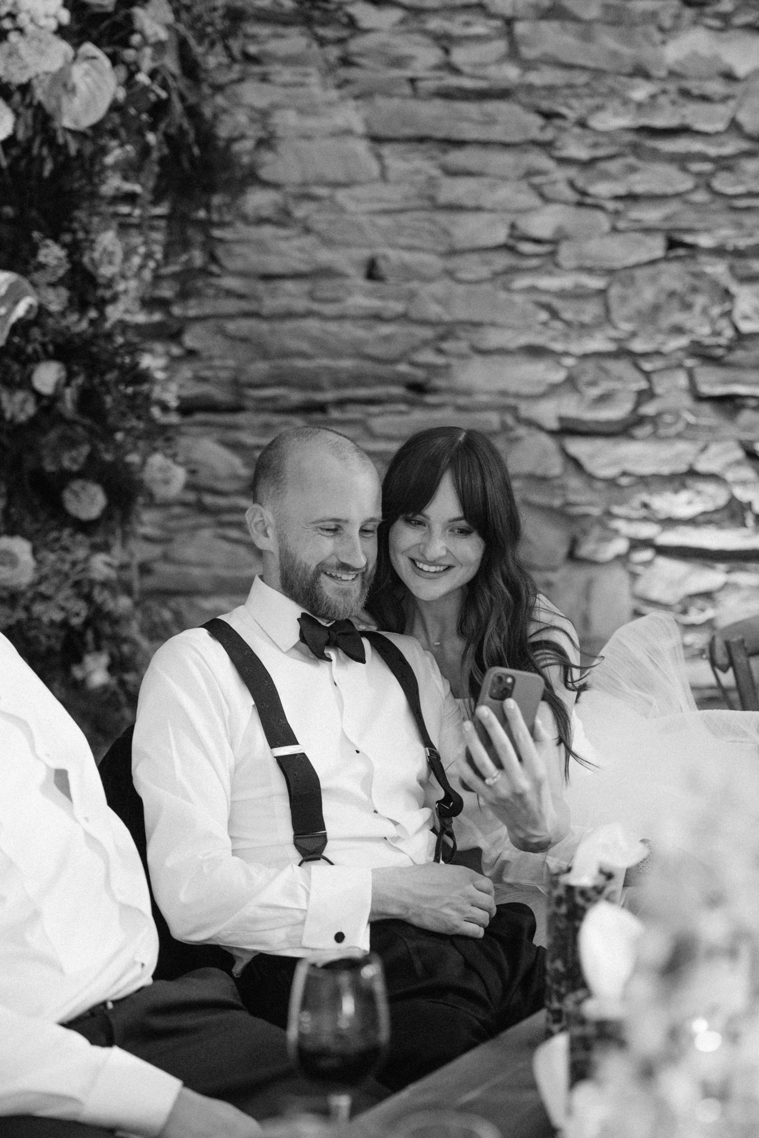 Wedding party members sharing joyful moments against a rustic stone wall backdrop.