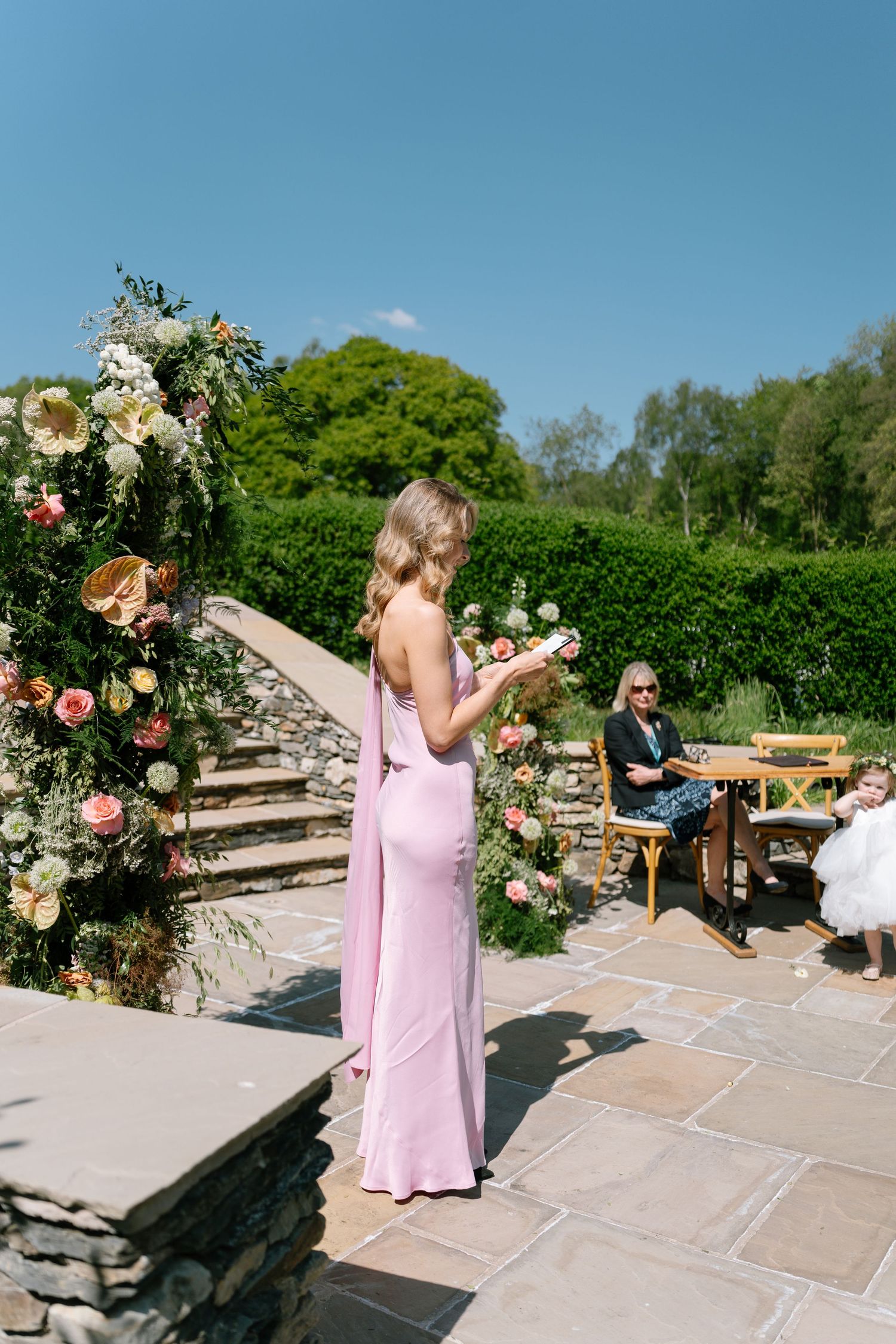 Wedding party members in pink dresses dancing on a sunny stone patio with floral decorations.