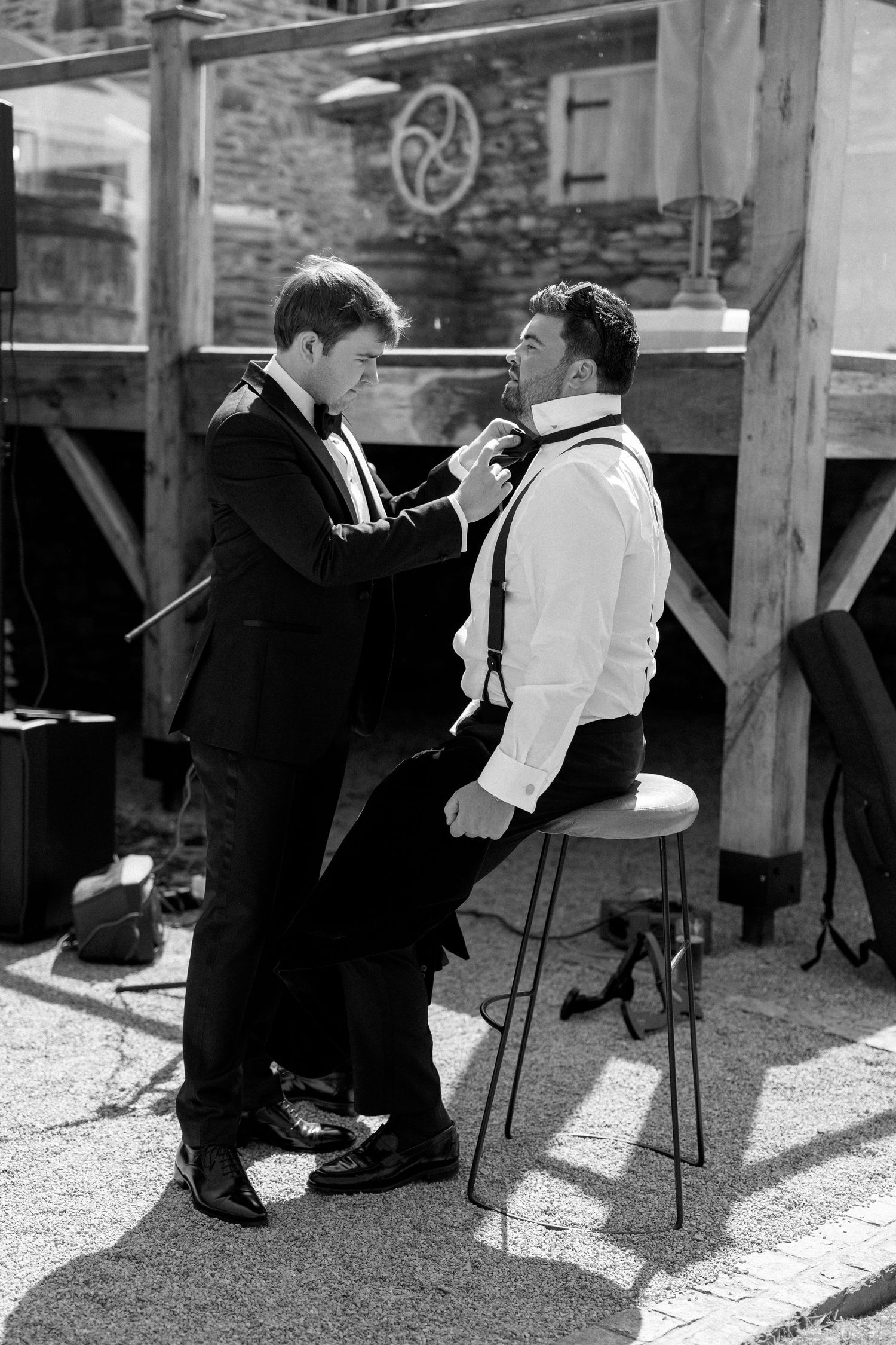 Two people in formal attire adjusting a bowtie during wedding preparations in black and white.