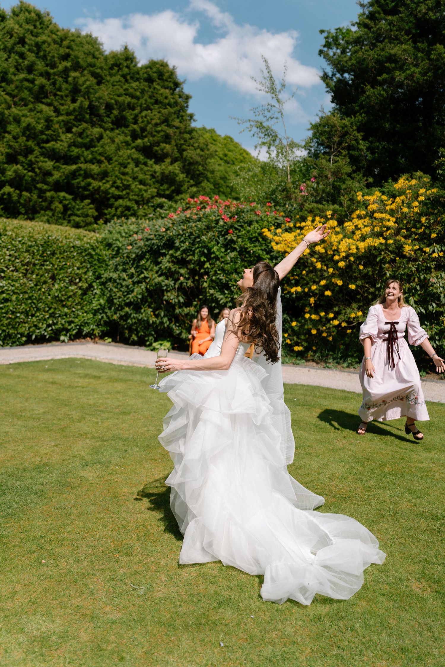 A bride in a white gown spins playfully on a sunny lawn with yellow flowers in the background.