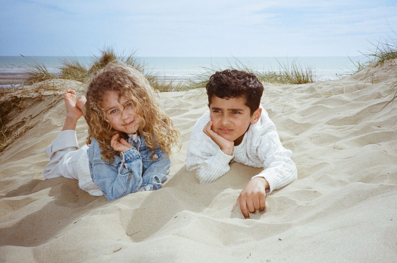 Two children lying on sandy beach dunes with ocean in background on a cloudy day.