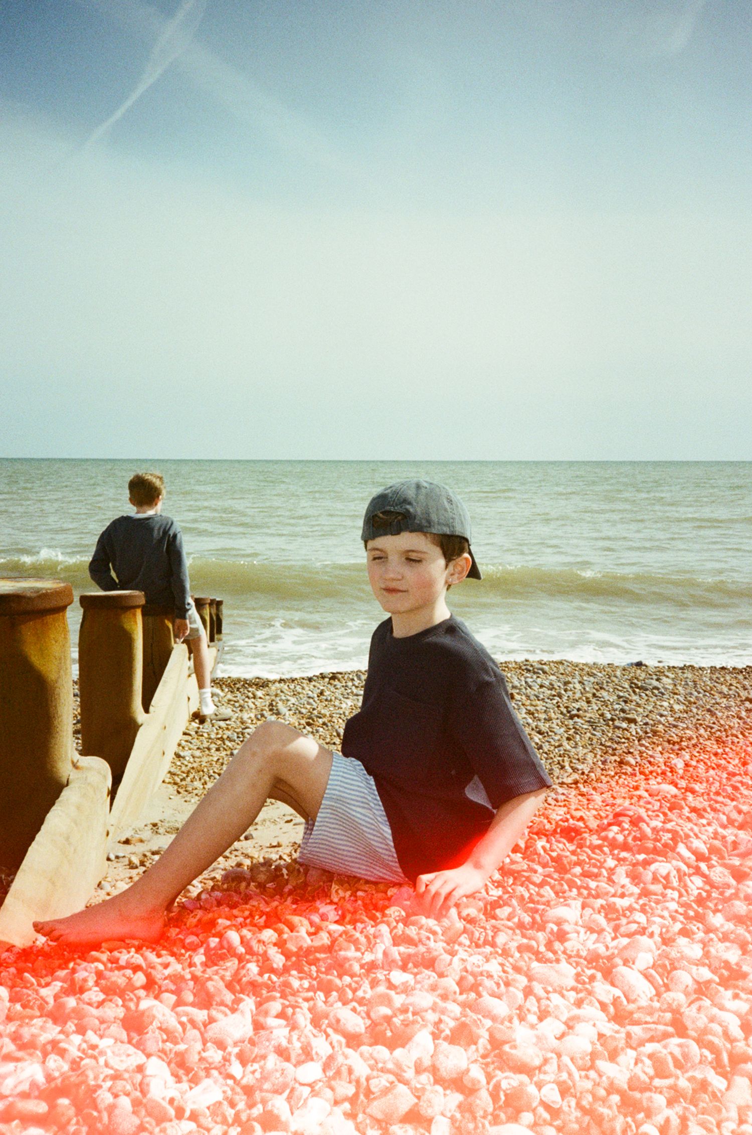 Boy in cap sits on pebble beach with red light leak effect, another person stands by groynes near calm sea.