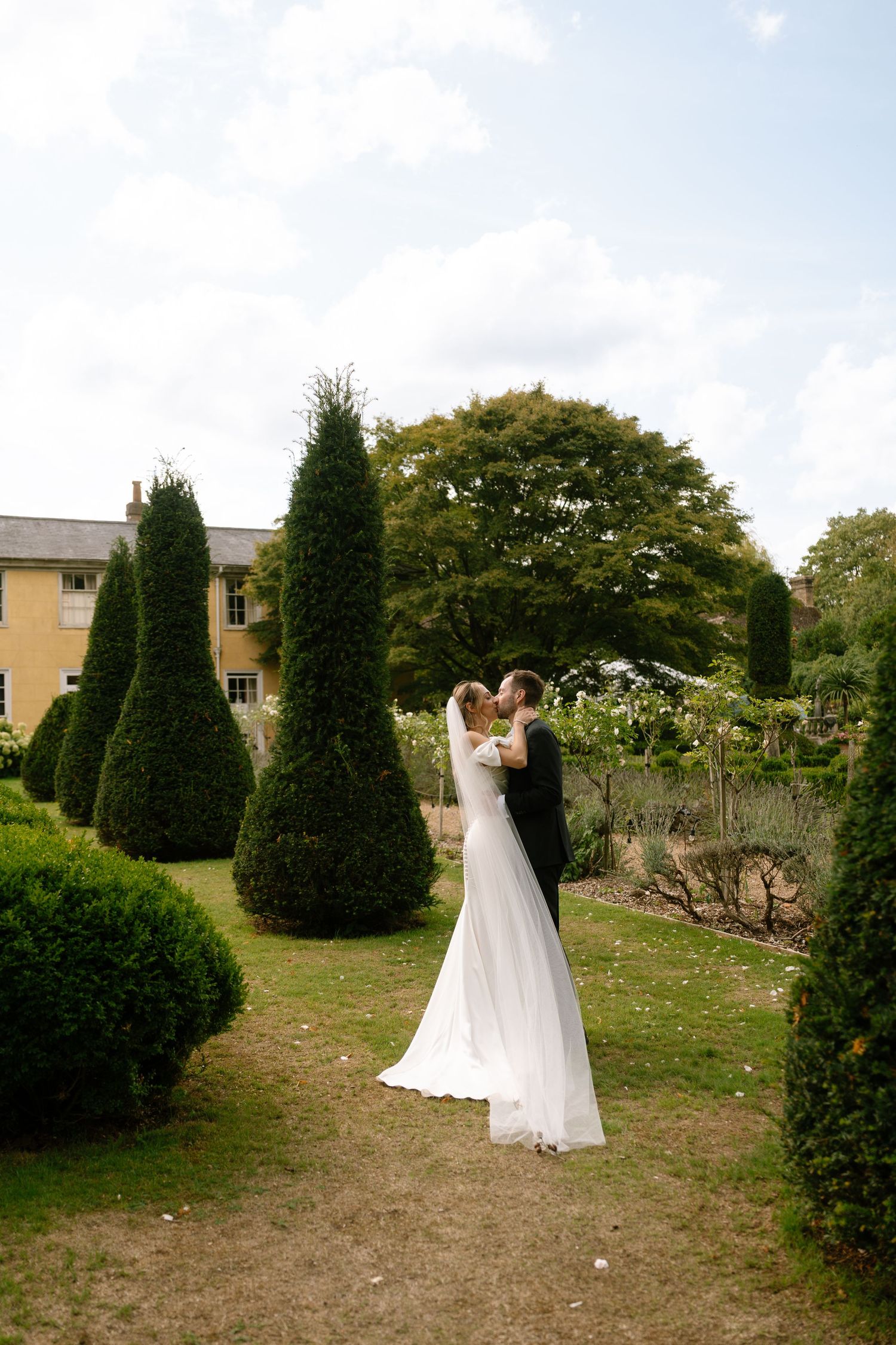 Wedding couple poses among manicured topiary trees in an elegant garden setting.