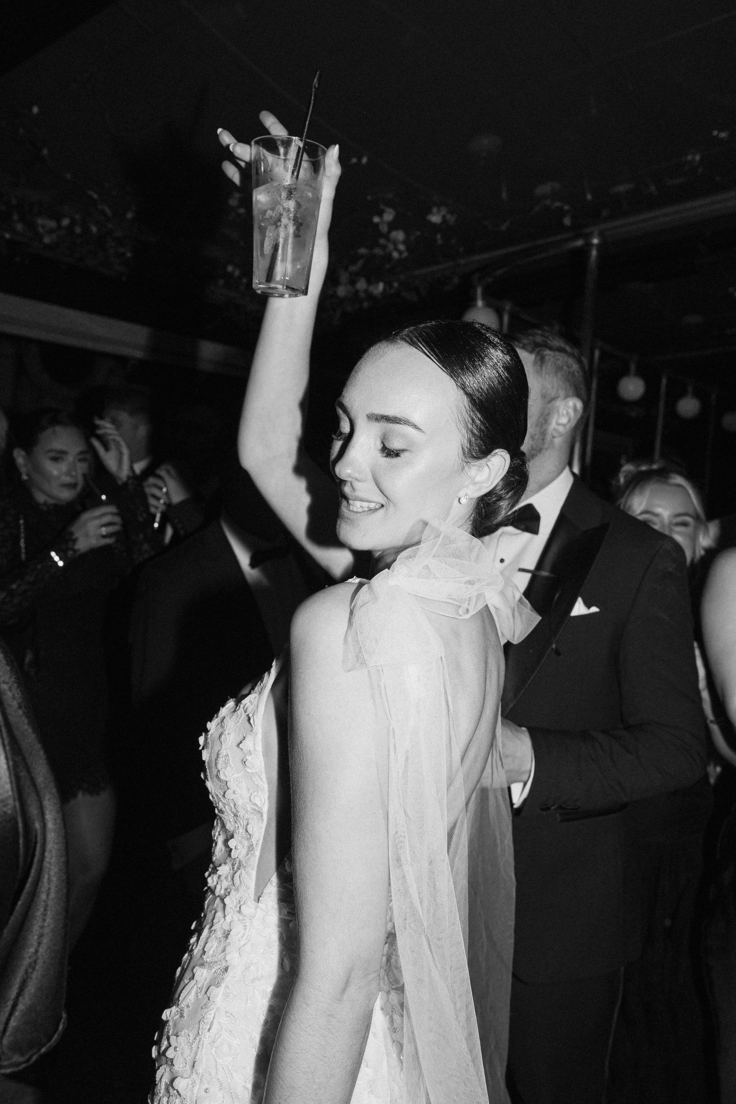 Wedding party dancing with raised arms in a dimly lit venue.