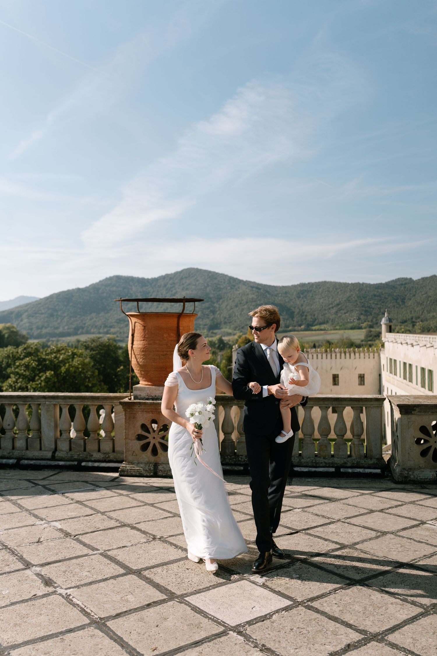 Wedding party poses on sunny castle terrace with mountain views and large decorative urn.