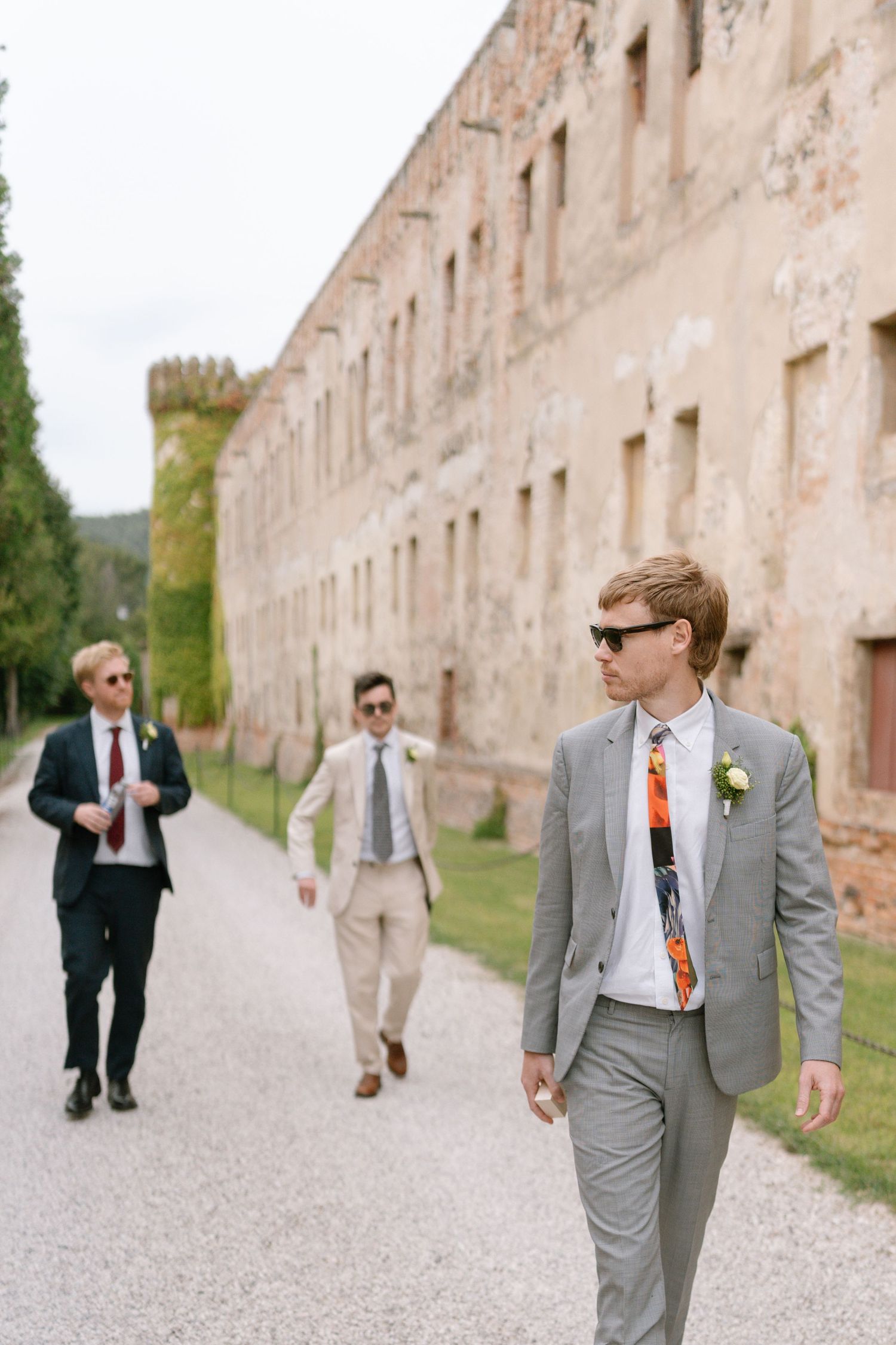 Wedding party members in gray suits stroll along a stone wall on gravel path.
