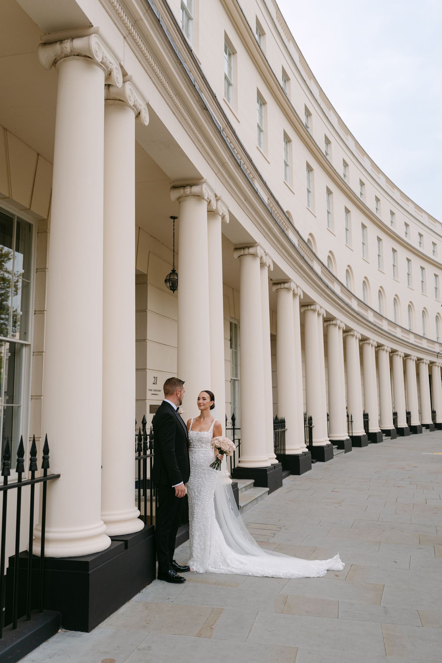 A wedding couple poses in front of a curved colonnade building with white pillars.