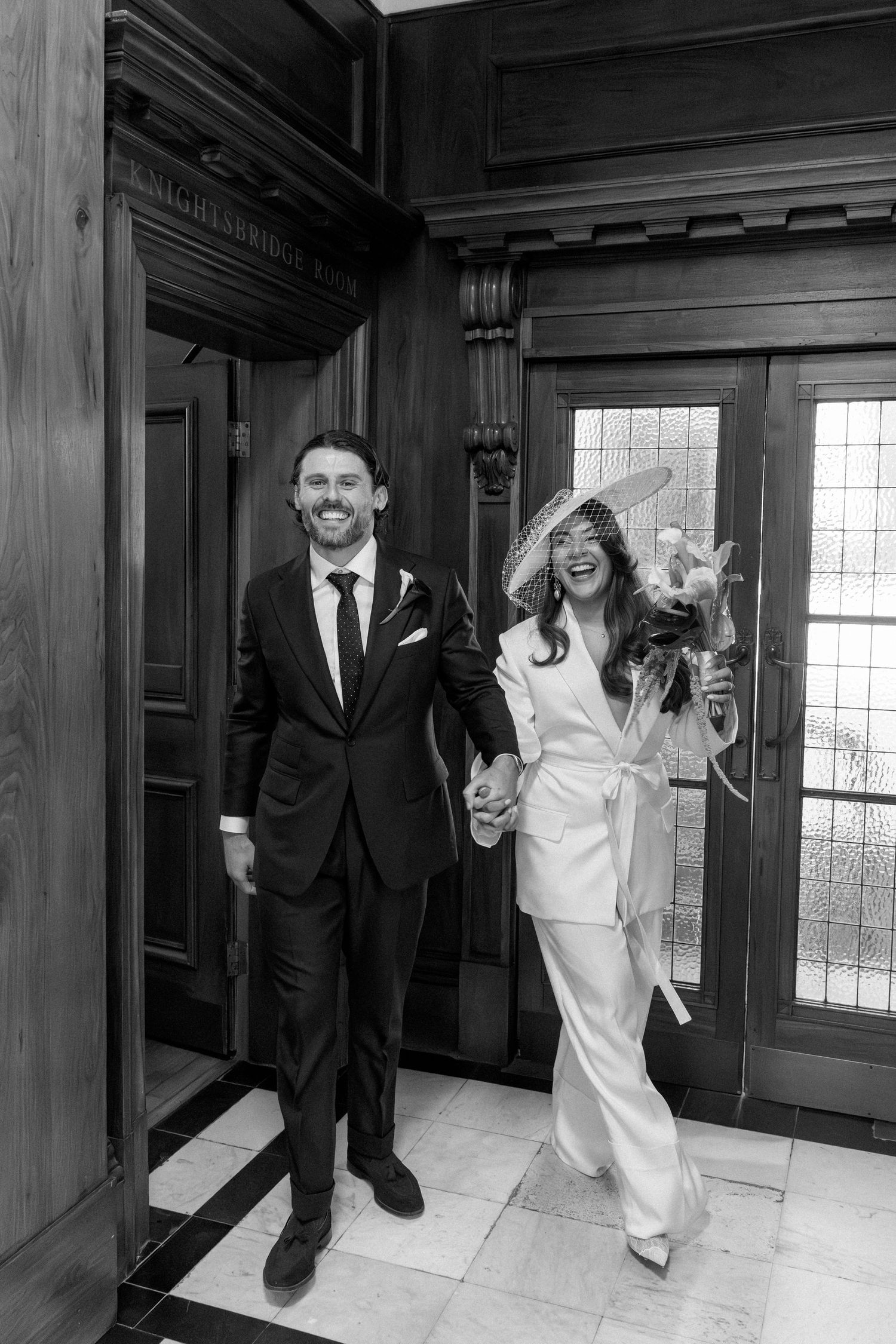 Black and white photo of smiling bride in white jumpsuit and hat holding groom's hand exiting elegant wooden doors.
