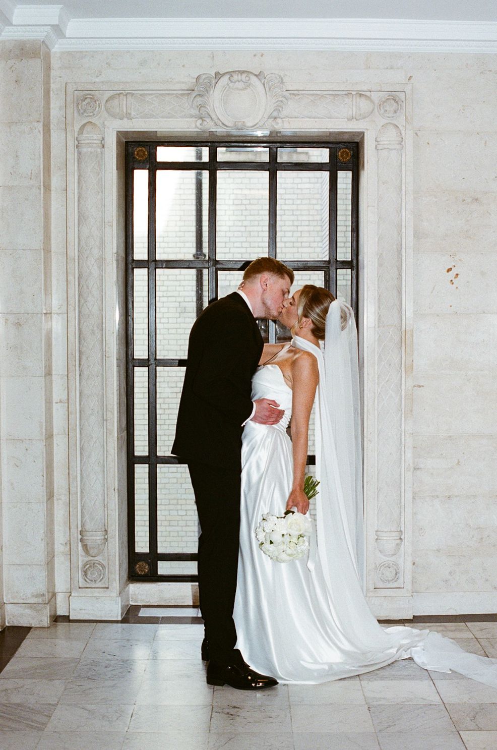 Bride and groom share a romantic kiss in front of an elegant marble doorway, bride holding white bouquet.