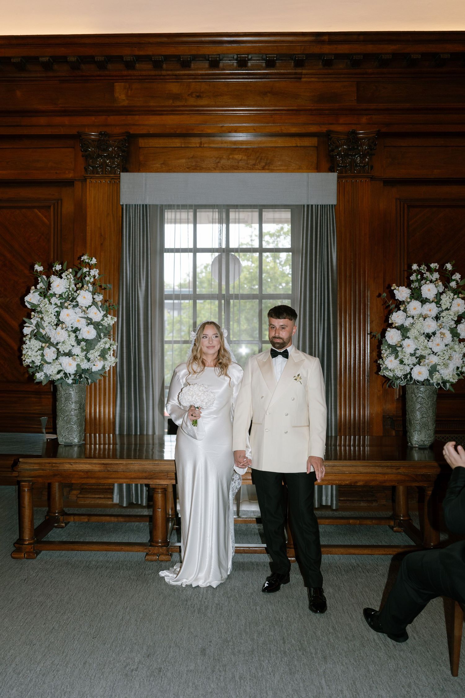 Wedding couple poses in an elegant wood-paneled room with white floral arrangements.